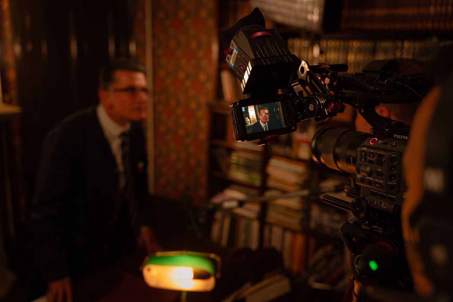 A TV camera films a man in a suit seated in a dimly lit room lined with bookshelves during an interview.