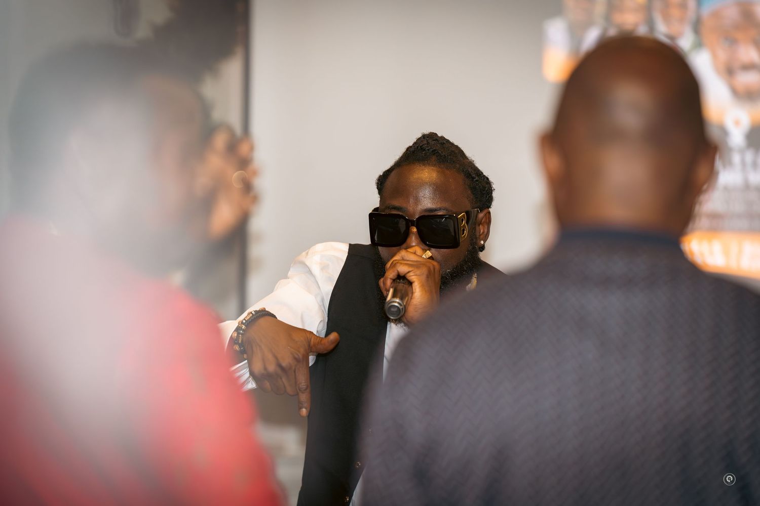 A person in sunglasses and a dark jacket sits at a bar with blurred patrons in the background.