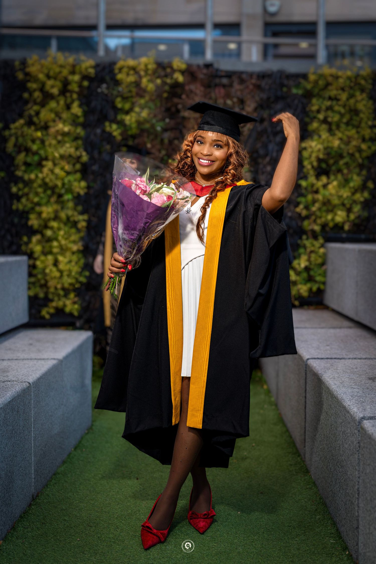 Graduate in black cap and gown with gold stole poses outdoors for graduation photos on concrete steps with greenery.