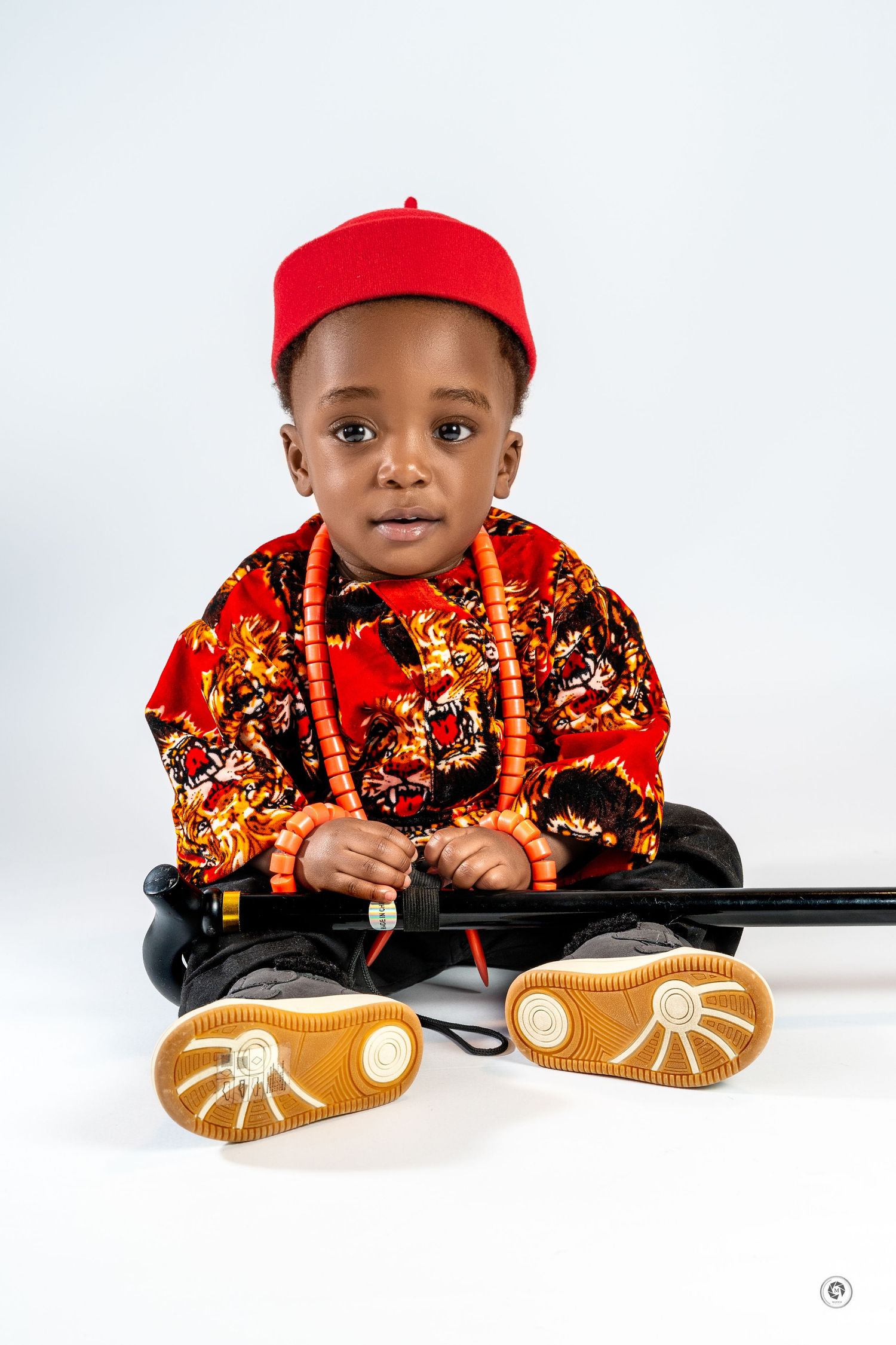 A small child in a red cap and floral jacket leans on a dark surface with a playful expression.
