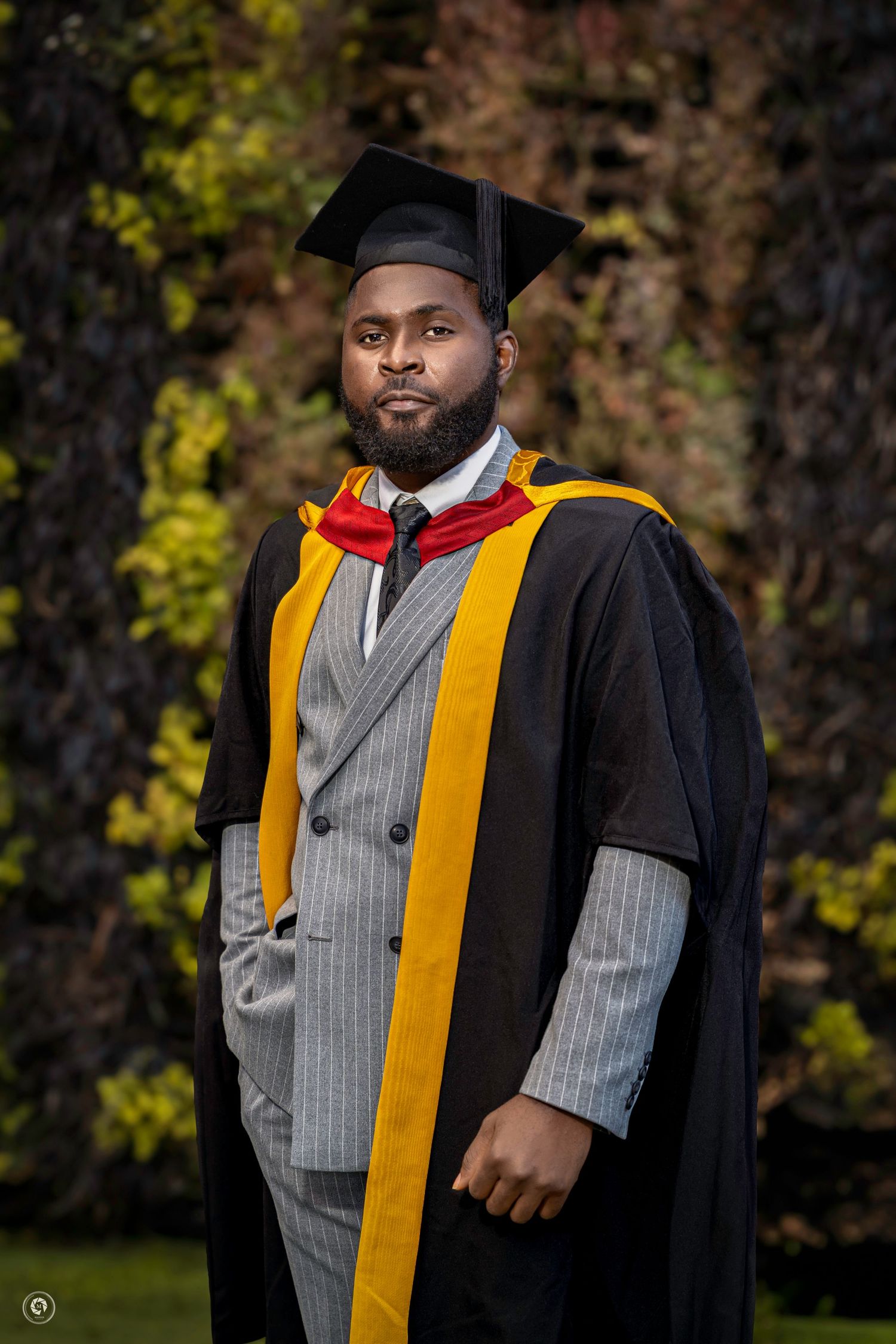 Graduate wearing black academic regalia with gold stole and gray vest poses outdoors against autumn foliage.