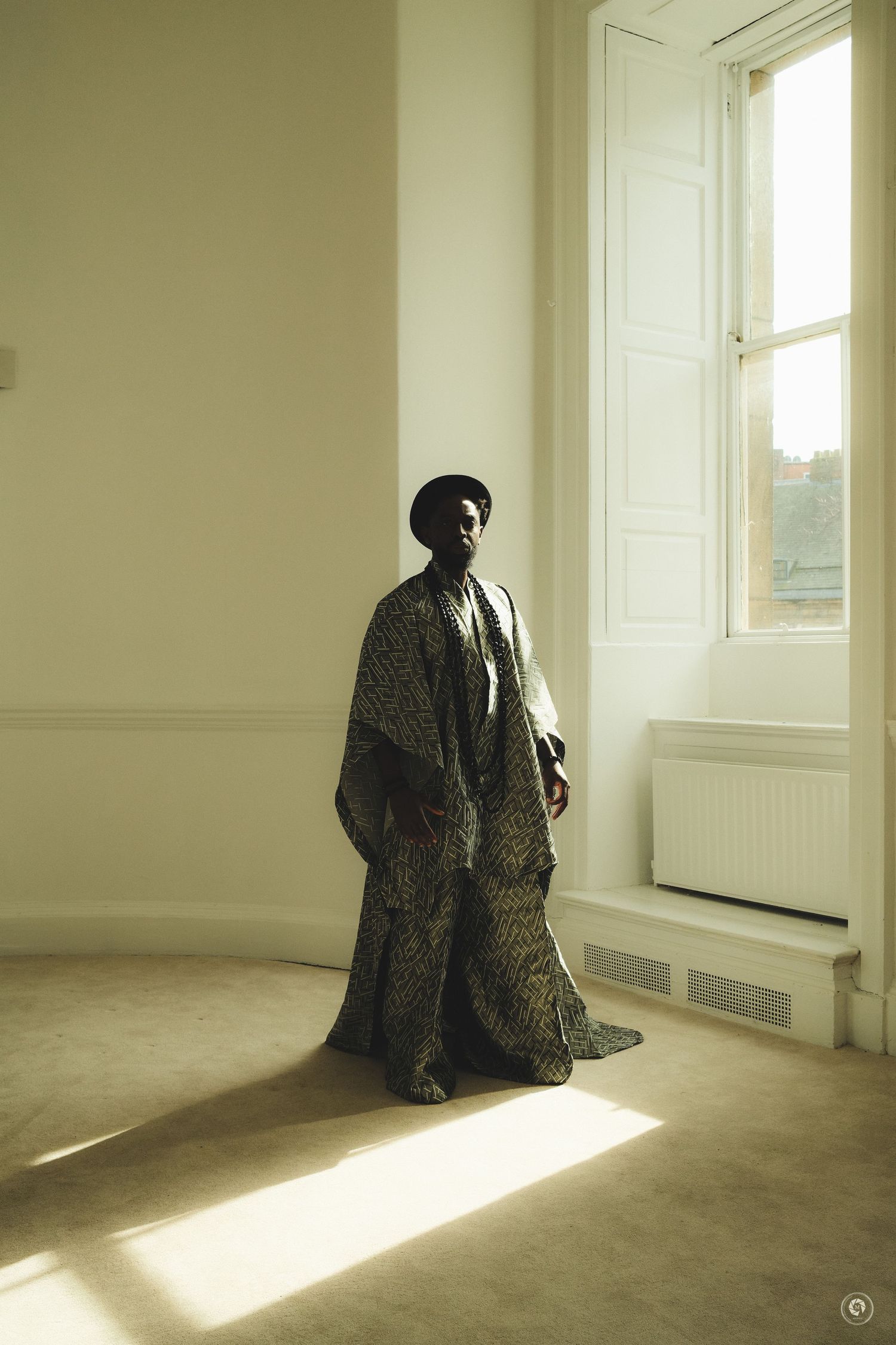Person in flowing patterned robe and beret standing in sunlit minimalist room near tall white window.
