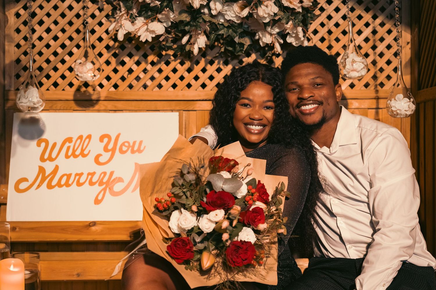 A romantic marriage proposal scene with illuminated signs reading 'Will You Marry' and floral decorations in warm lighting.