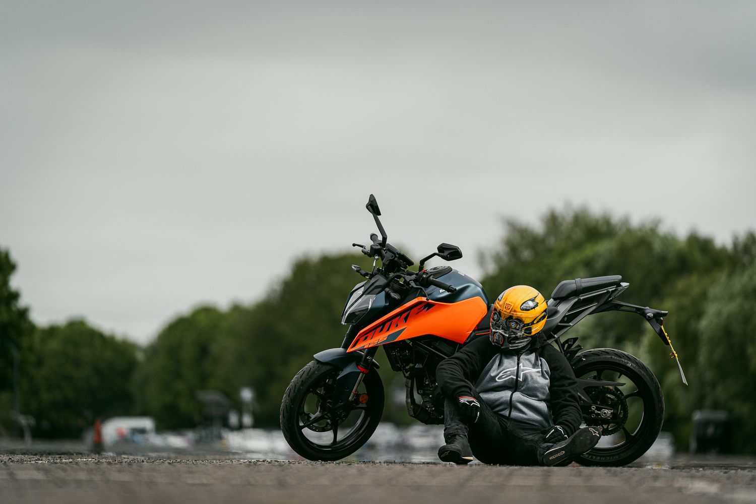 An orange and black KTM motorcycle parked on wet pavement with trees in the background on an overcast day.
