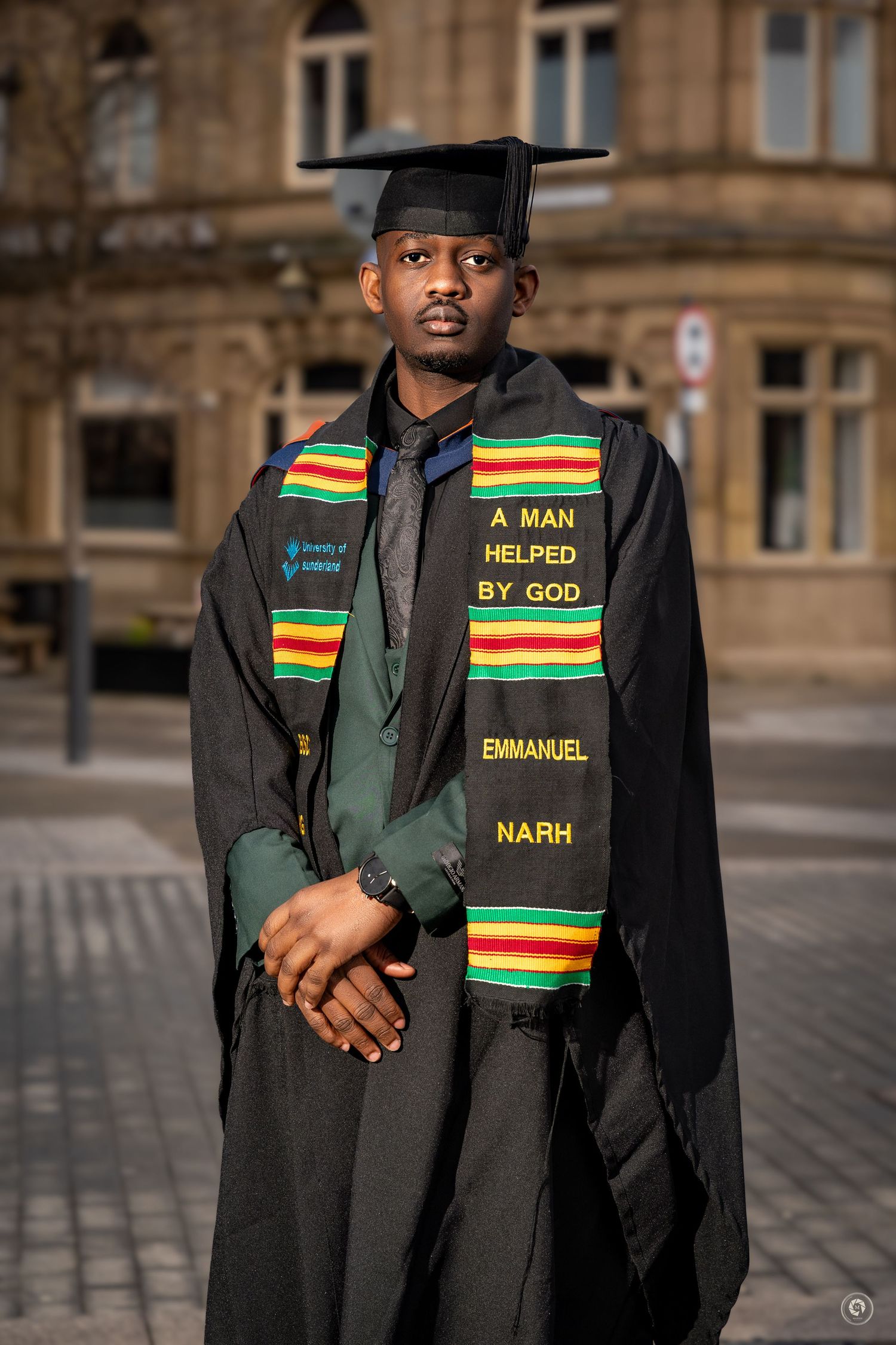 Graduate wearing black academic regalia with a colorful kente stole featuring text stands outside a stone building.
