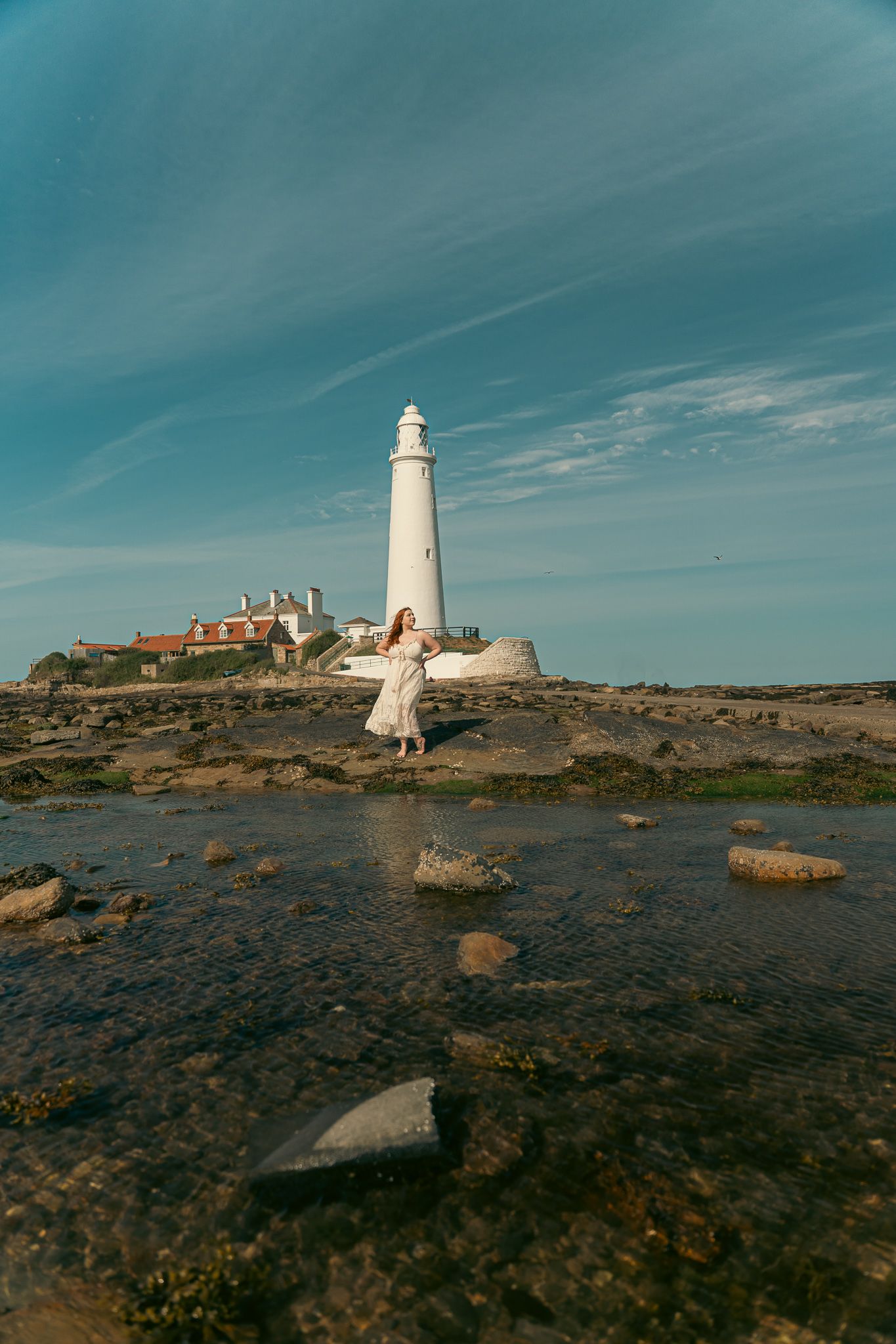 White lighthouse stands atop rocky coastal terrain against vibrant turquoise sky during golden hour.