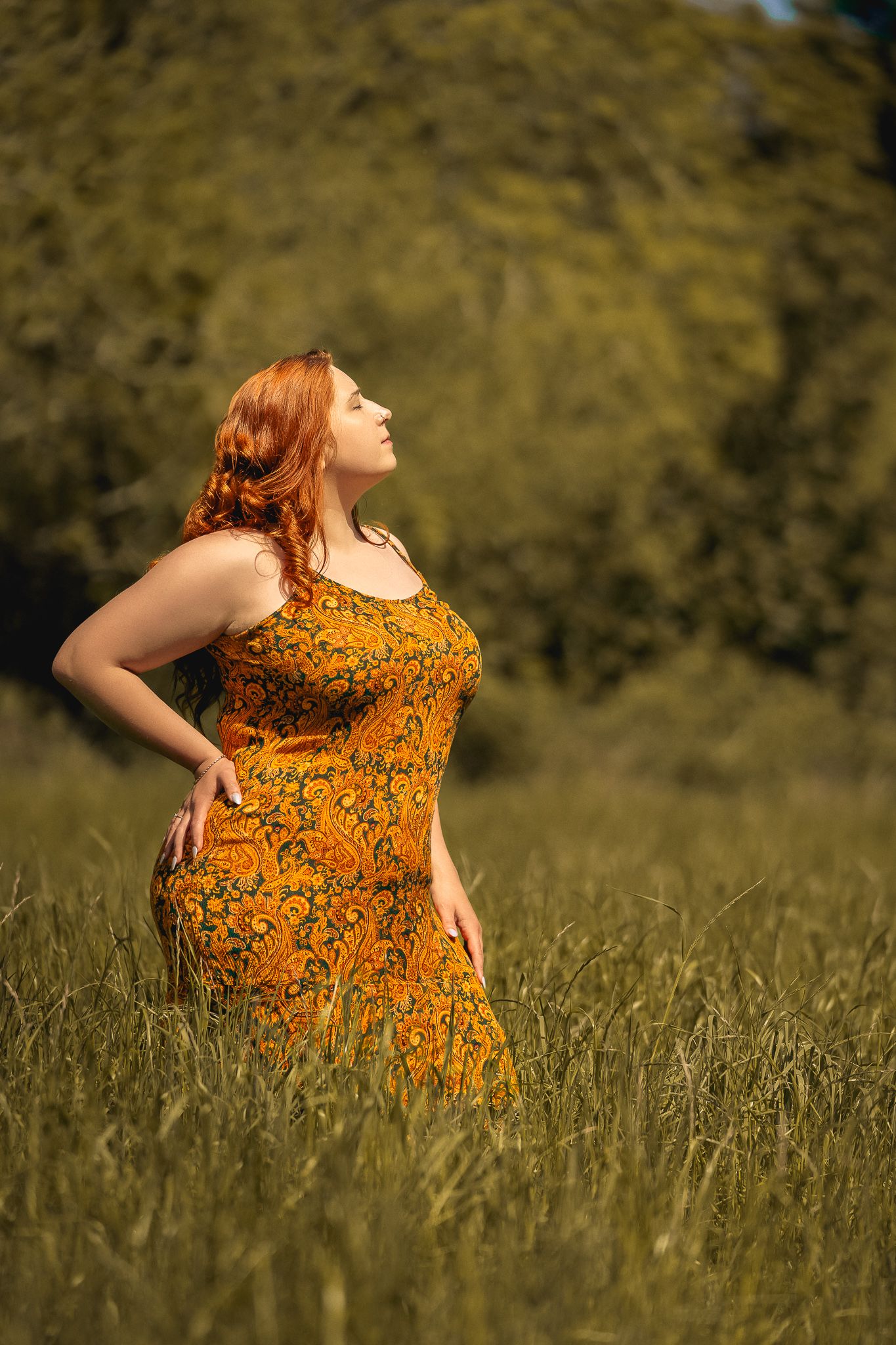 A woman in a vibrant orange floral sundress poses gracefully in a sunny meadow during golden hour.