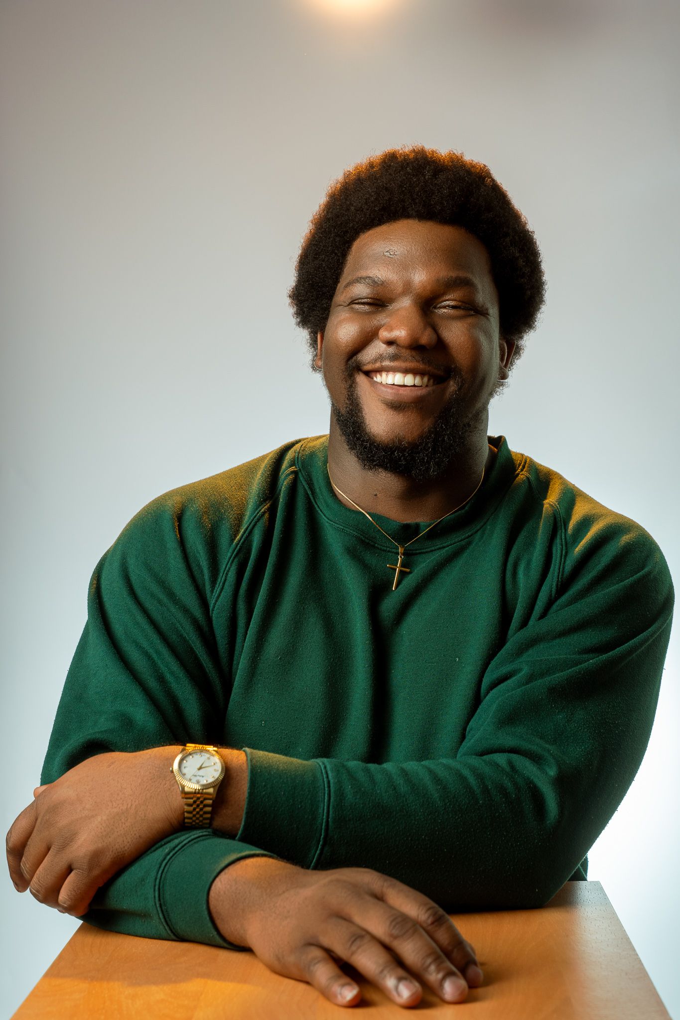 A person wearing a dark green sweater smiles warmly while sitting at a wooden table wearing a gold watch and cross necklace.