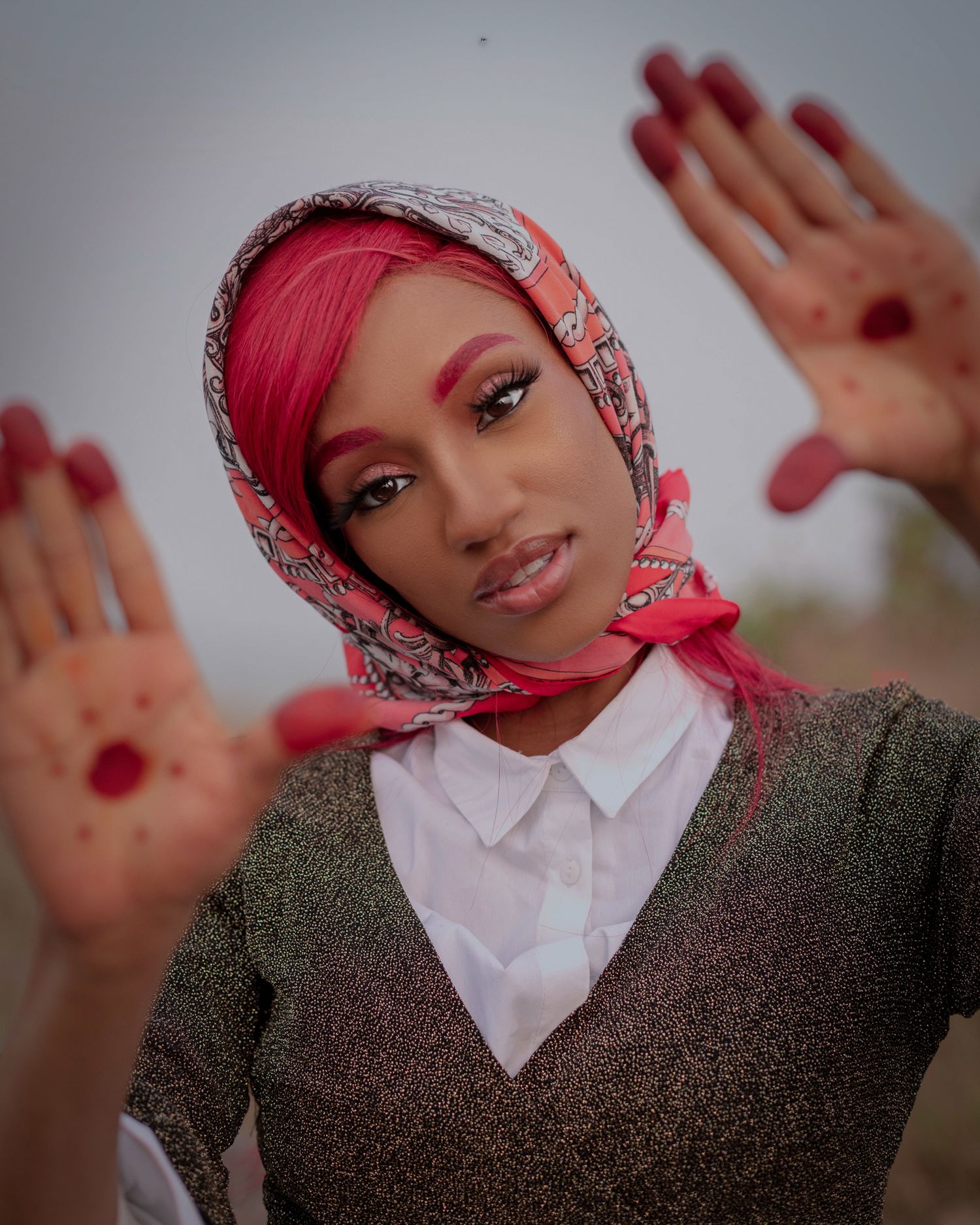 Wearing a patterned headscarf and pink makeup, a person in a gray sweater presses their hands against glass with painted nails.