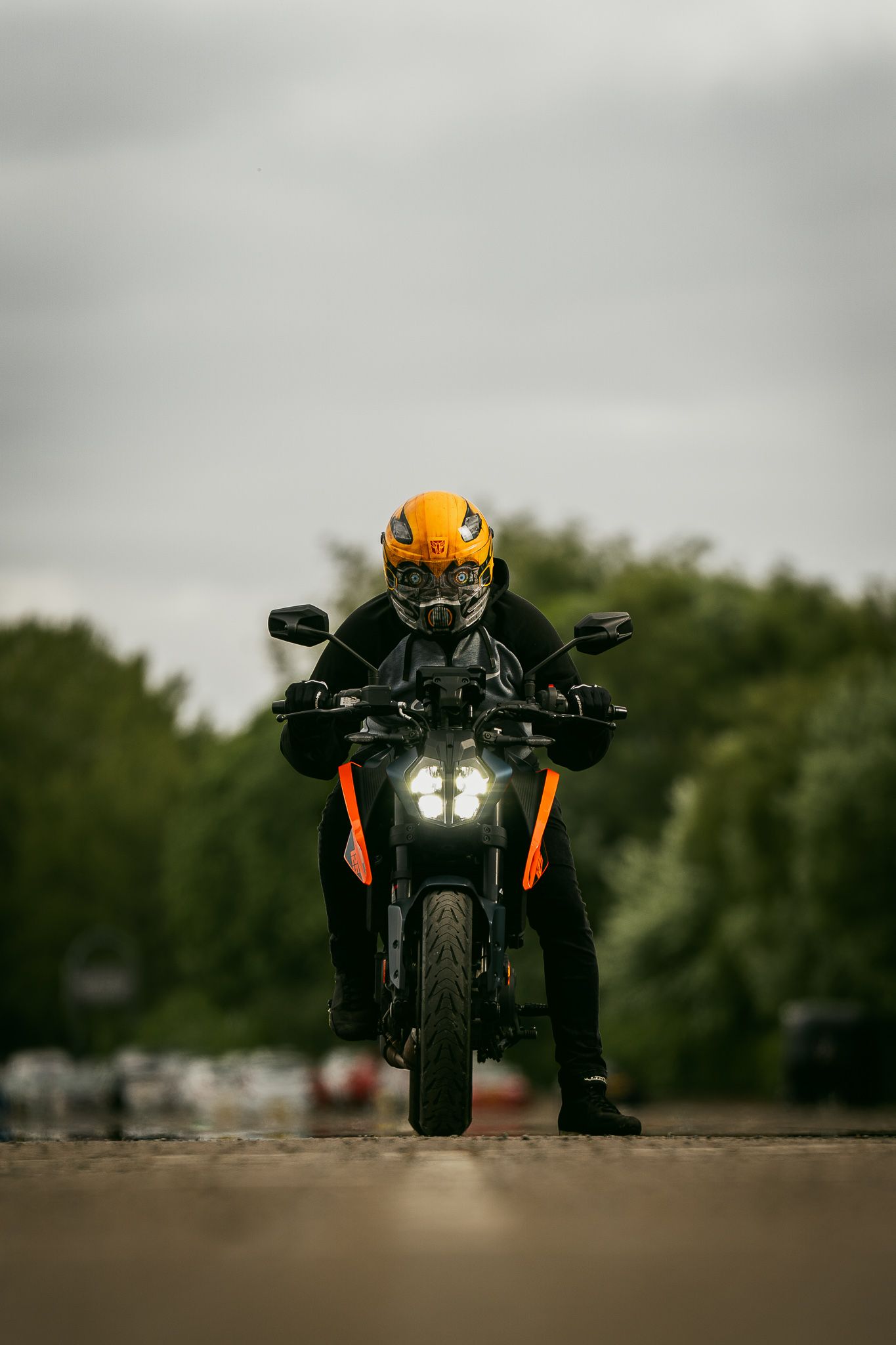 Motorcyclist wearing orange helmet rides black sports bike on asphalt road with headlight illuminated on overcast day.