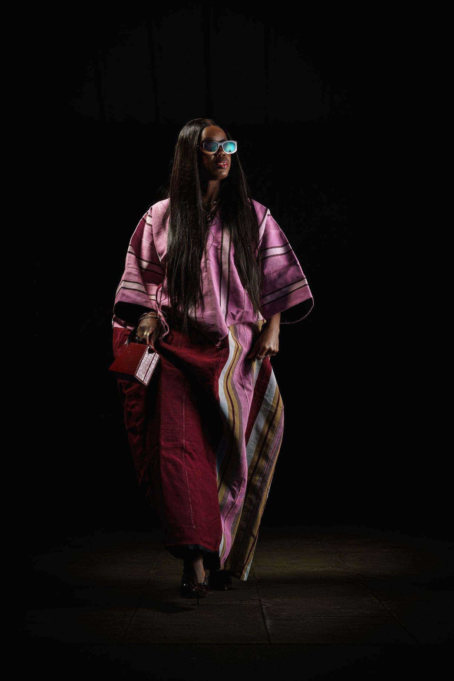 A model walks the runway in a flowing pink and red striped kaftan dress with blue sunglasses and a red handbag.