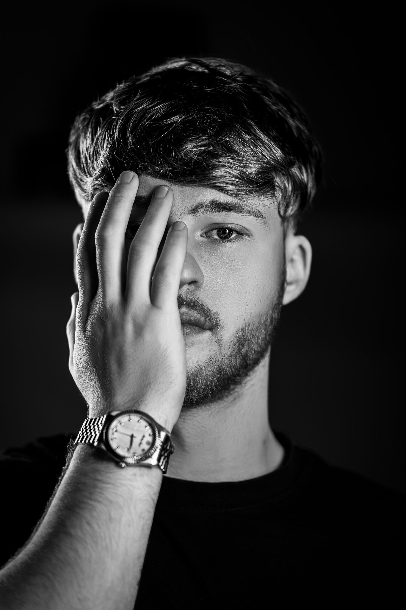 Black and white artistic portrait of a person covering half their face with a watch-adorned hand against dark background.
