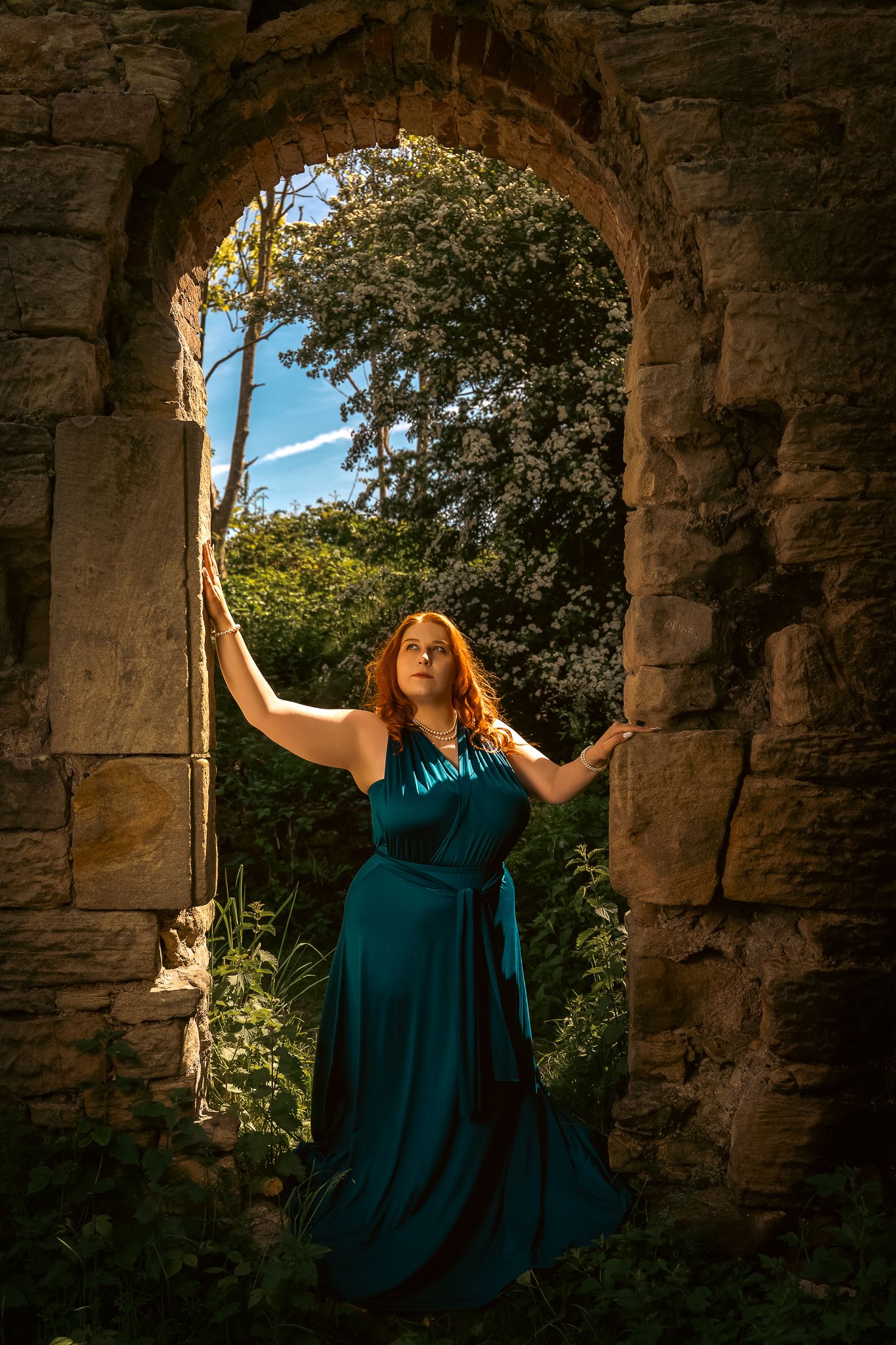 A person in a teal gown poses dramatically in an ancient stone archway with sunlight streaming through the opening.