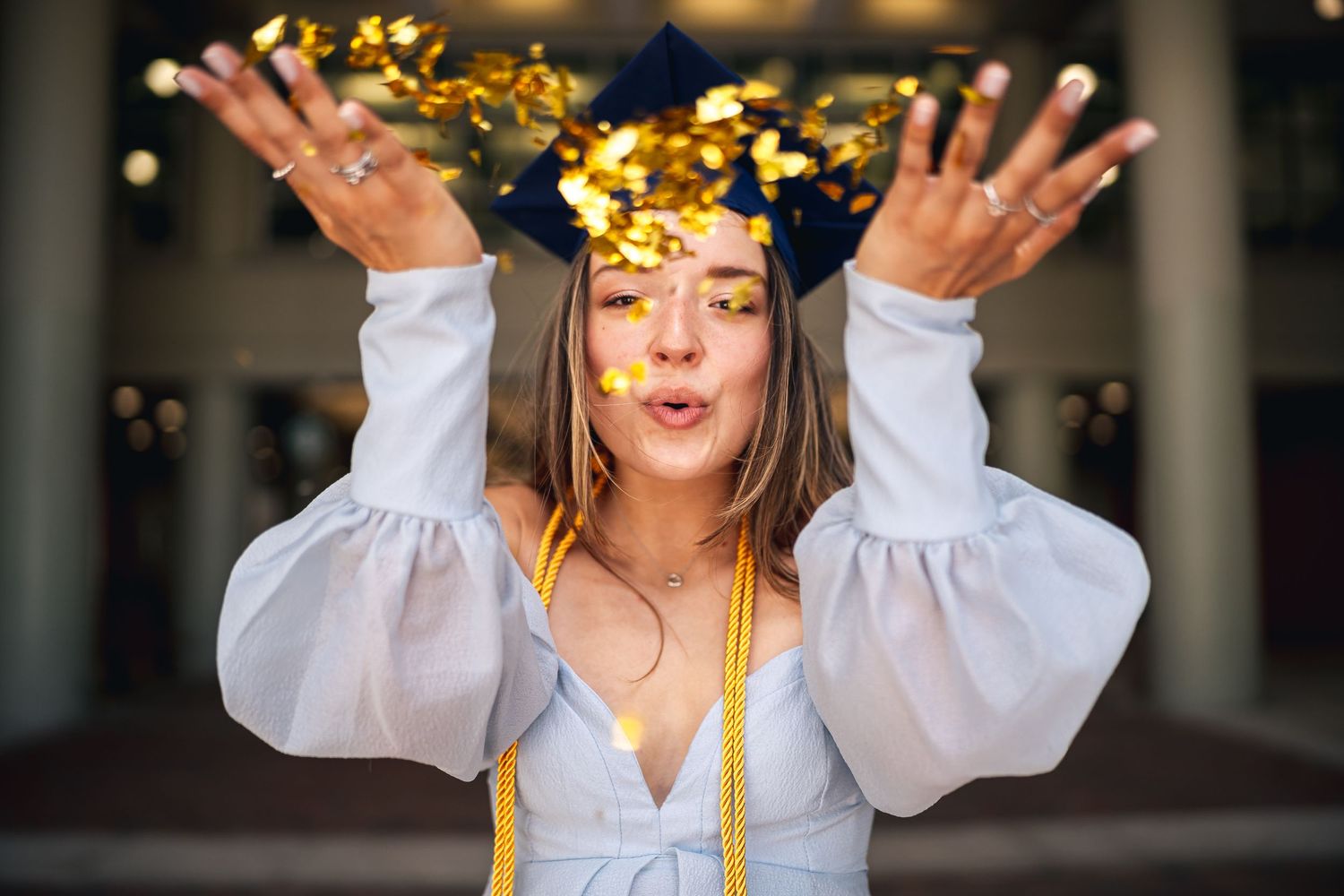 Graduate celebrating by tossing gold confetti while wearing white graduation gown and cap.