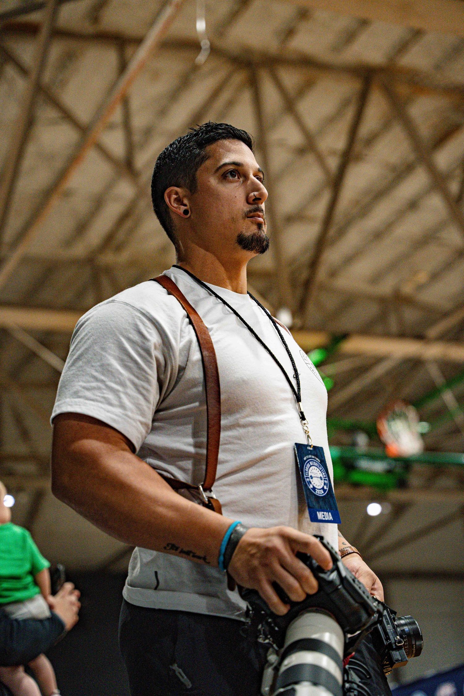 Professional photographer in white shirt and suspenders holding camera with telephoto lens at indoor event.
