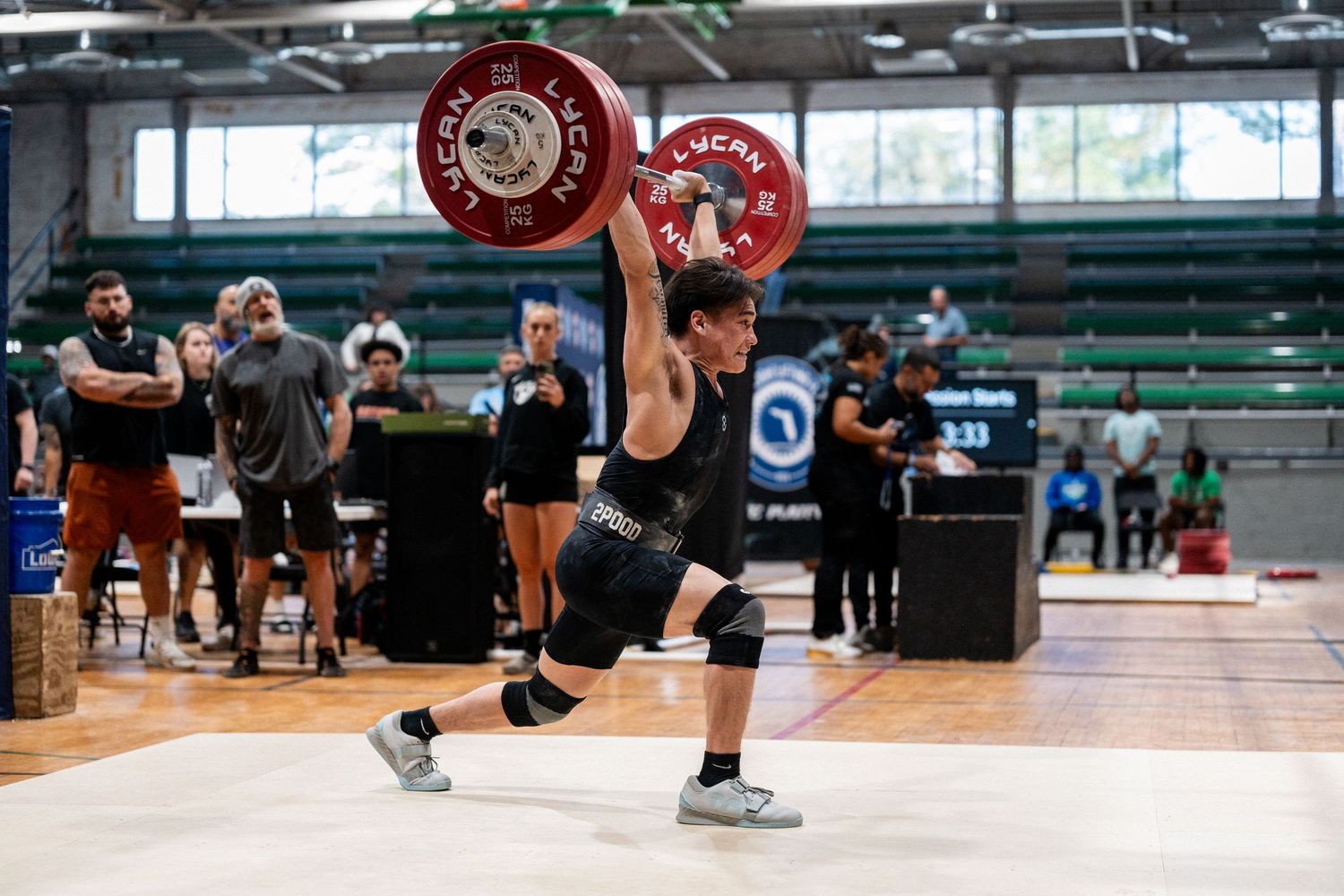 Weightlifter performing an overhead lift with red plates during indoor competition while spectators watch in background.