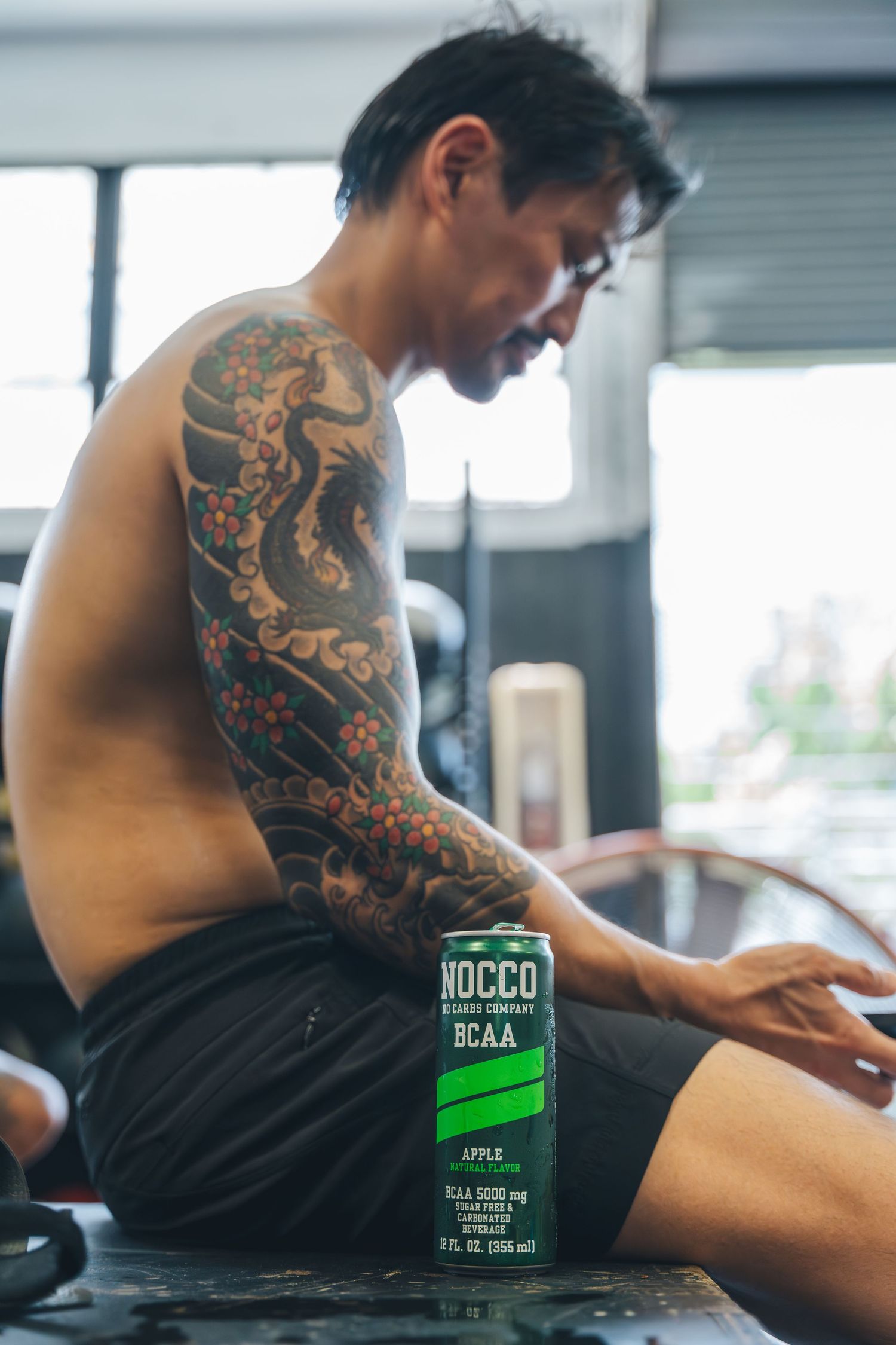 Person with intricate sleeve tattoo sits shirtless next to a Nocco BCAA drink bottle.