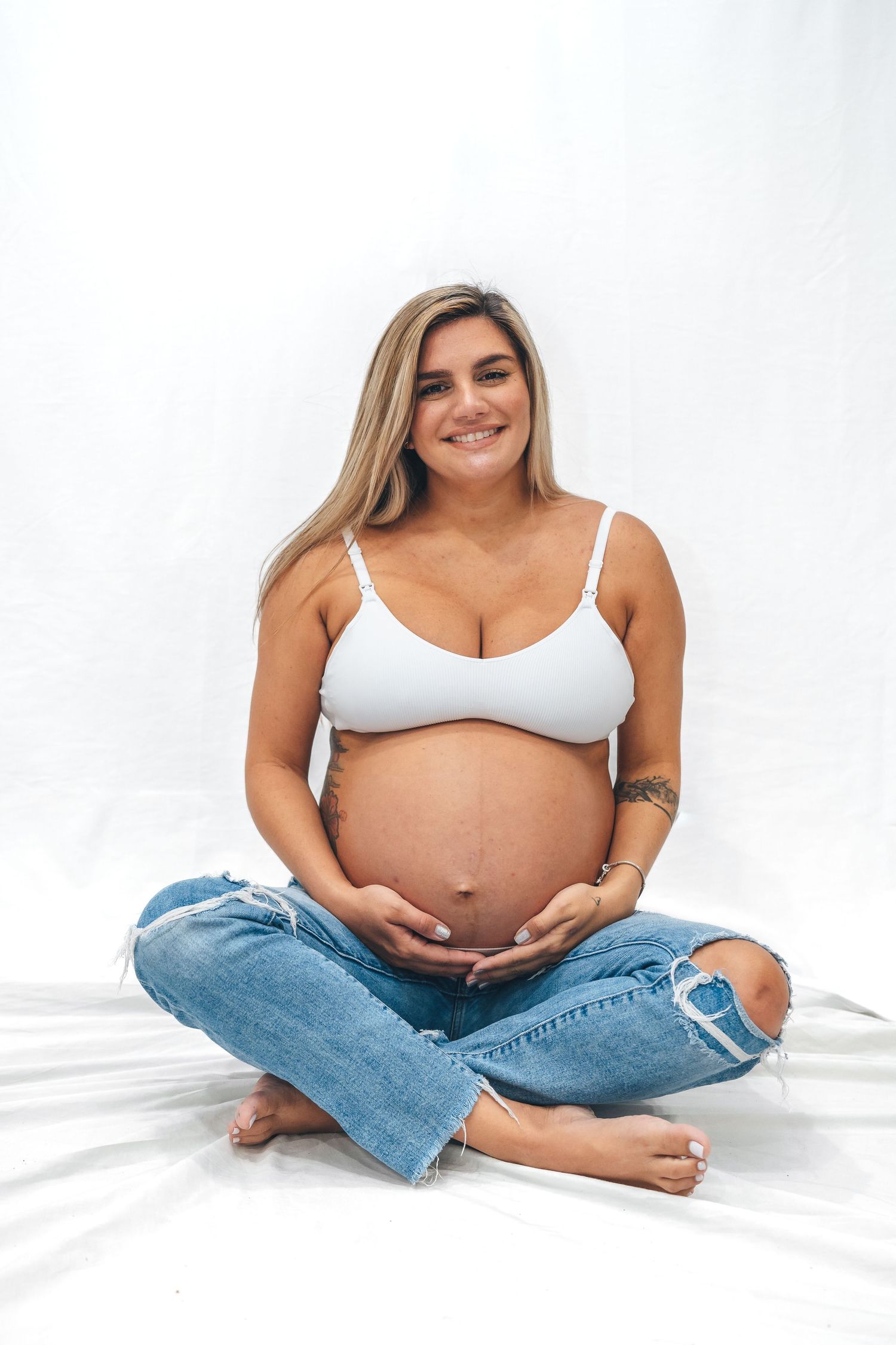 Expectant mother in white top and ripped jeans sits cross-legged on white background.