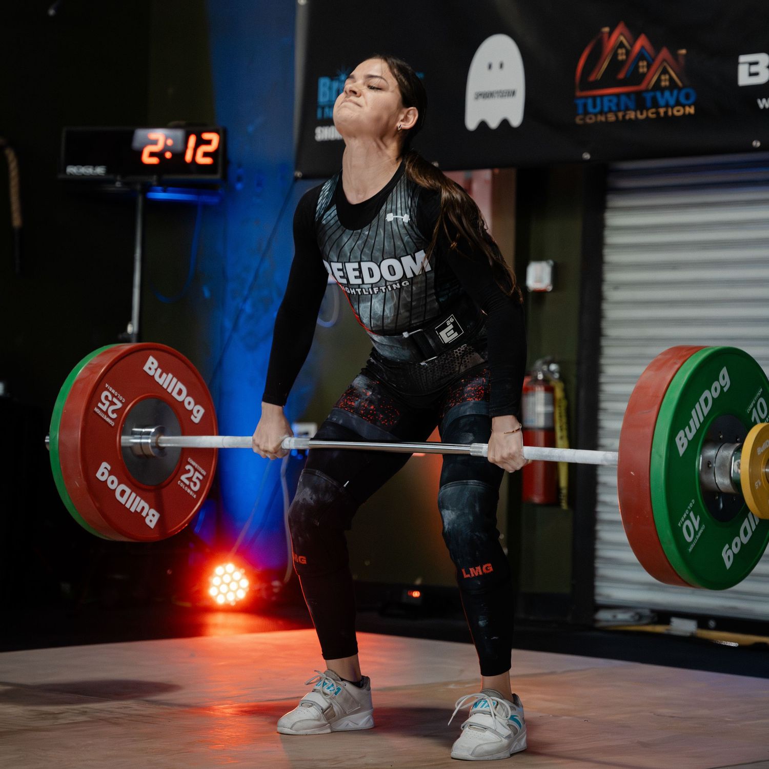 An athlete performs a heavy weightlifting exercise in a gym with colored weight plates and digital clock display.