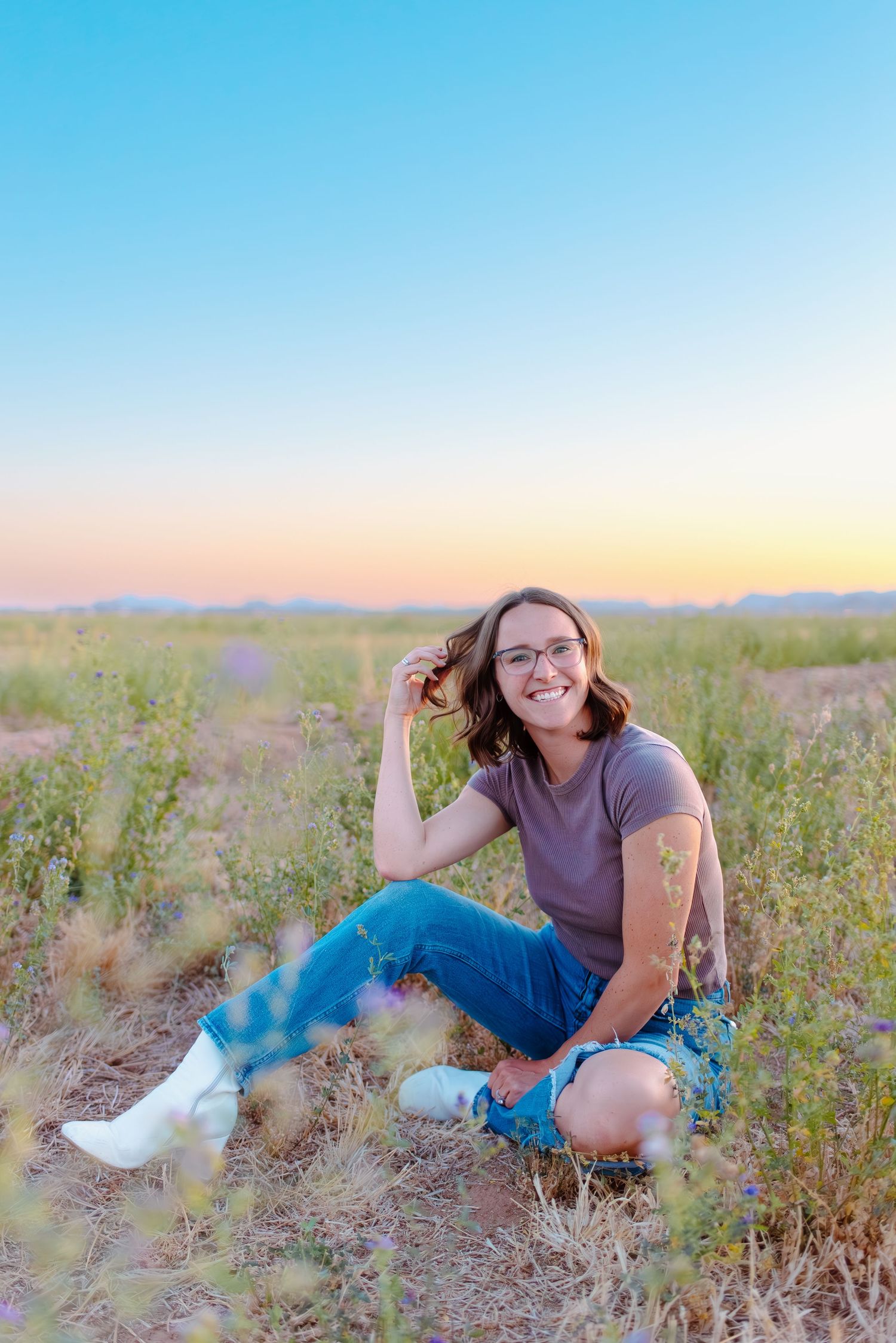Person sitting in a desert field at sunset wearing blue jeans, a purple shirt and white shoes.