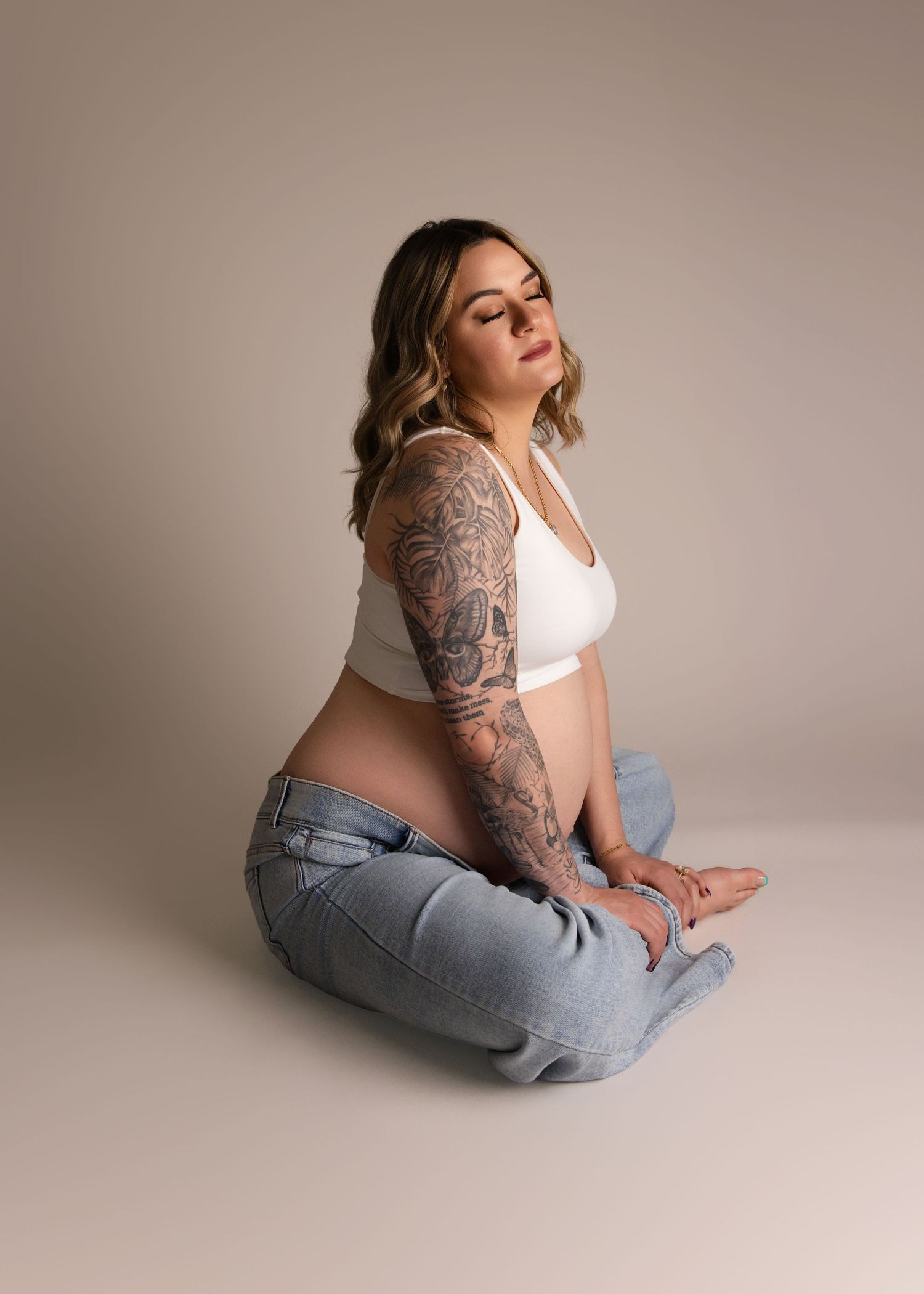 Portrait of someone in light jeans and white top sitting on floor with intricate sleeve tattoos in minimalist studio setting.