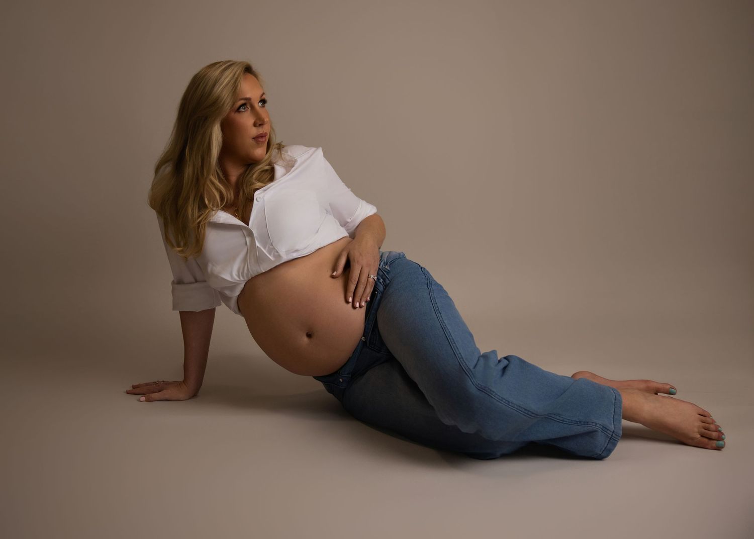 Pregnant person in white crop top and jeans reclines on floor during maternity photoshoot with neutral background.