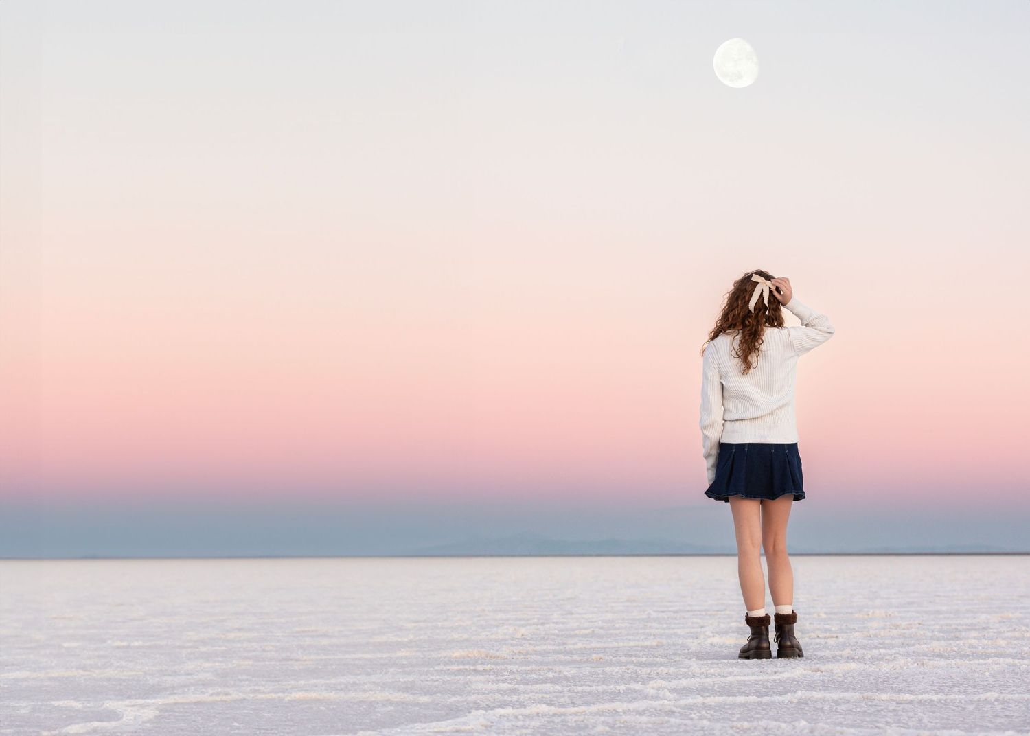 A person stands on the salt flats at sunset with a pink sky and full moon above.