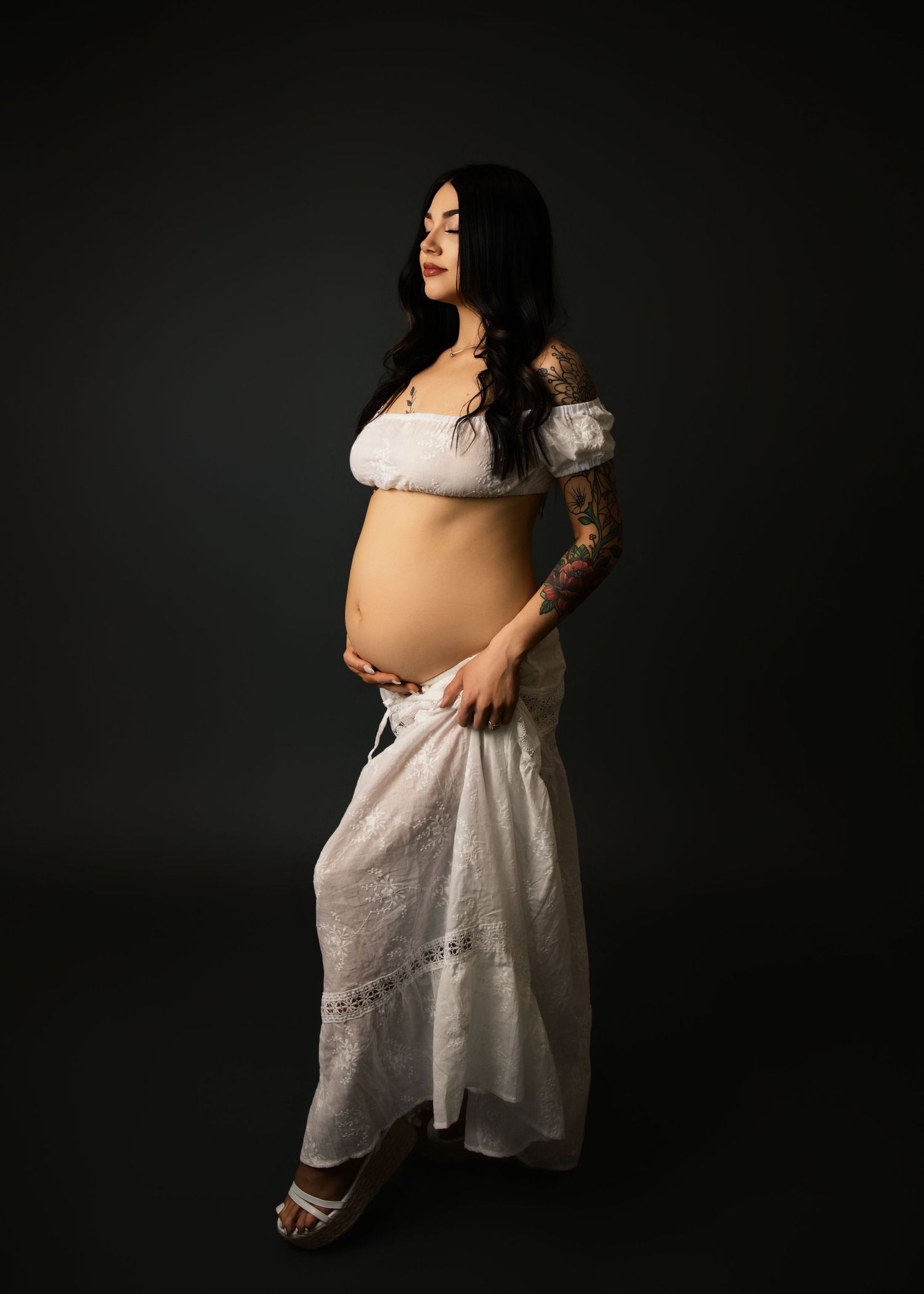 Artistic maternity portrait of an expectant mother wearing a flowing white dress against a dark background.