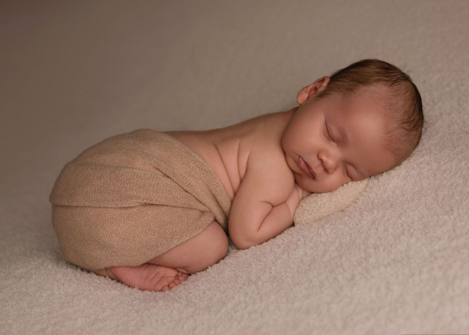 A peaceful newborn sleeps while curled in fetal position on soft beige blankets in a minimalist photography setup.