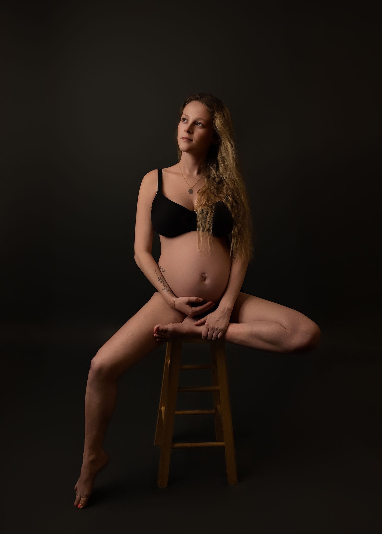 Expecting mother in black lingerie poses gracefully on wooden stool against dark studio backdrop in artistic maternity photographer photo.