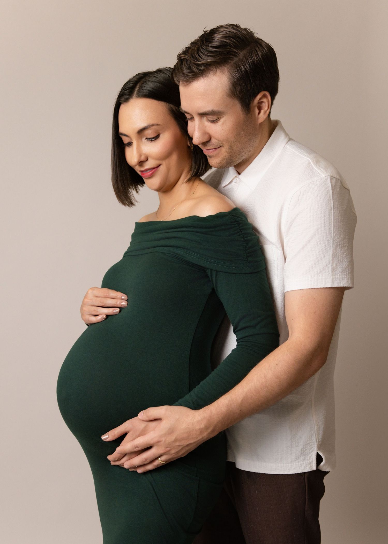 A couple in intimate maternity pose with woman wearing green off-shoulder dress and man in white shirt against beige backdrop.