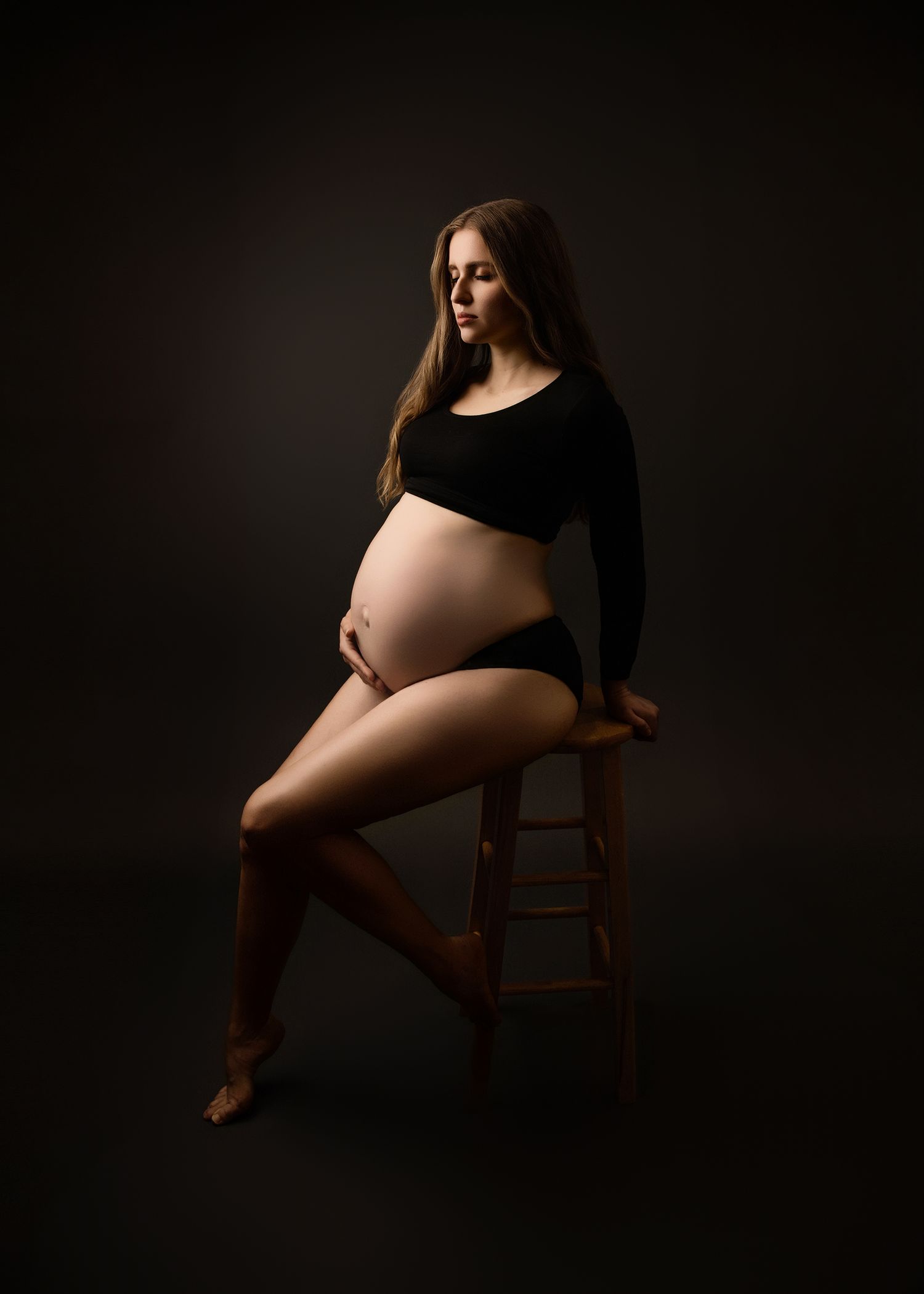 Artistic maternity photographer portrait of an expectant mother sitting on a wooden stool against a dark background with dramatic lighting.