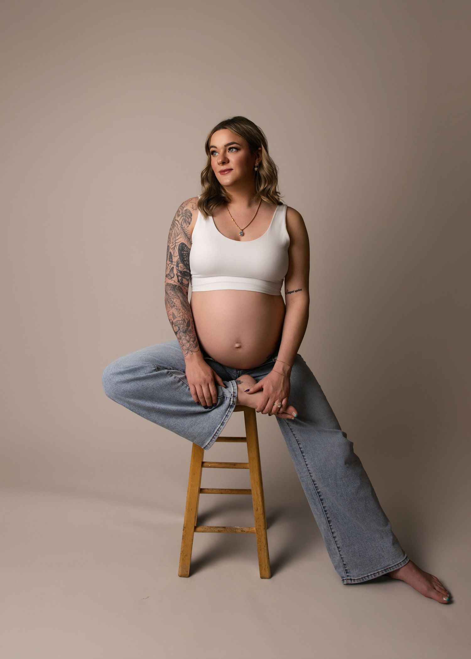 A pregnant person in a white sports bra and jeans sits on a wooden stool against a beige backdrop, showing arm tattoos.