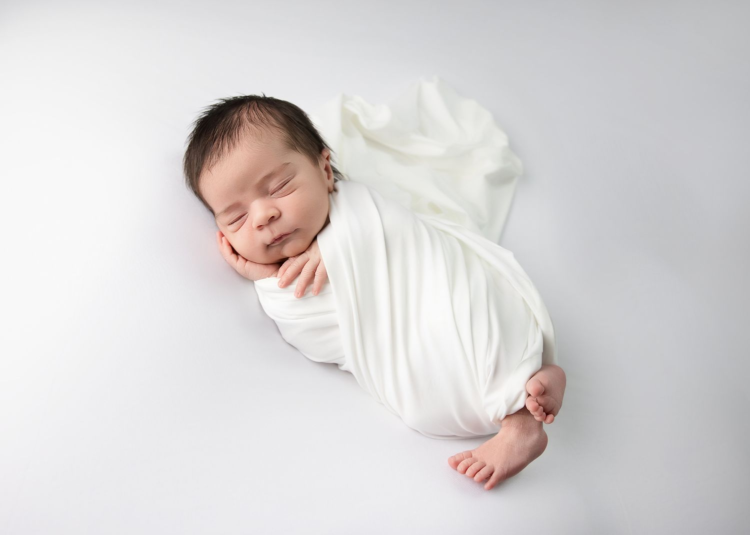 A peacefully sleeping newborn wrapped in soft white fabric lying on a light gray backdrop.