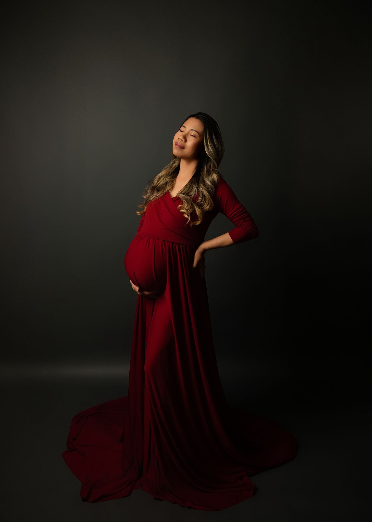 A maternity photographer takes photo of pregnant woman in a flowing burgundy velvet gown posing elegantly against a dark studio backdrop.