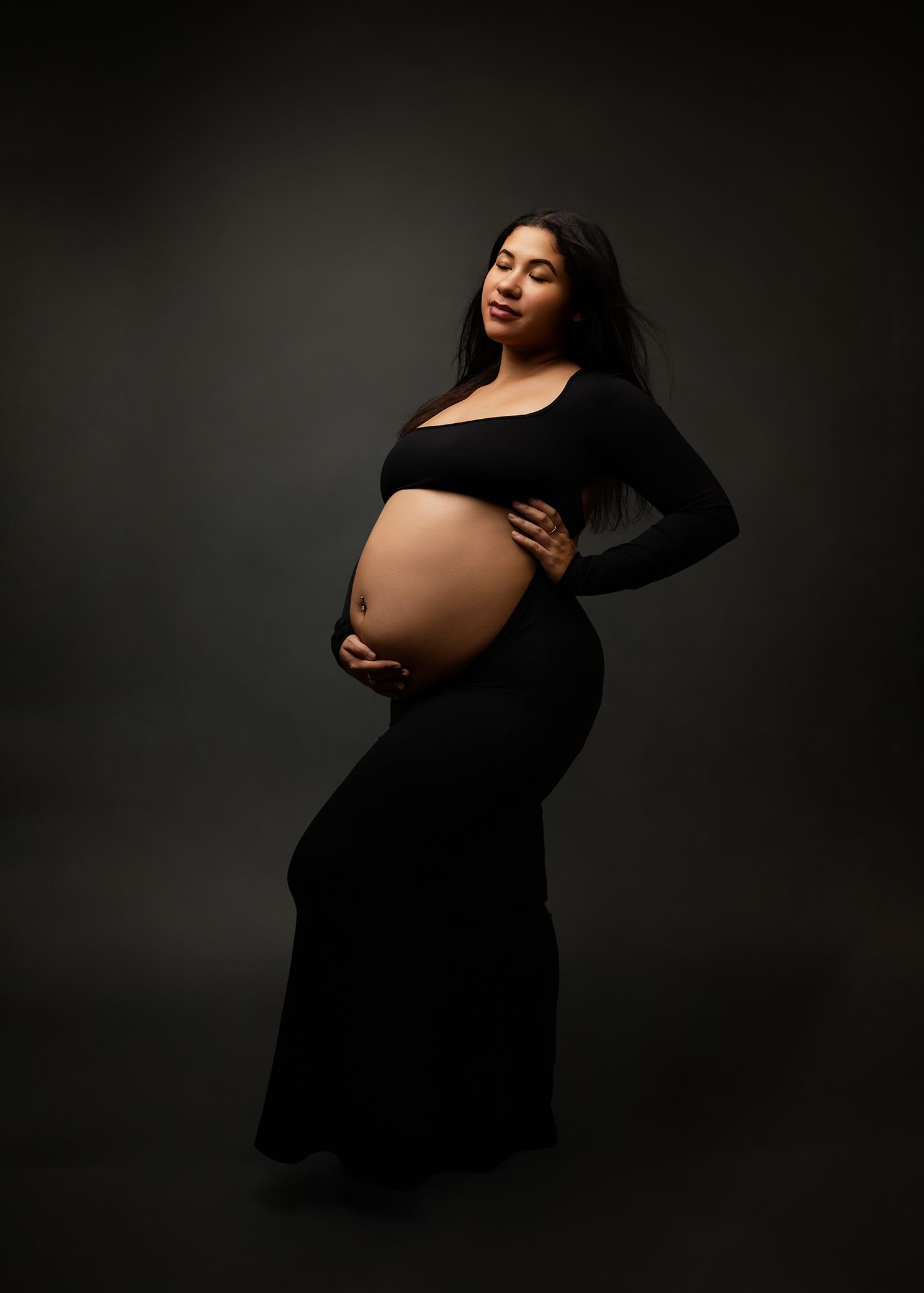A stunning maternity portrait of an expectant mother in a black dress posing elegantly against a dark background.