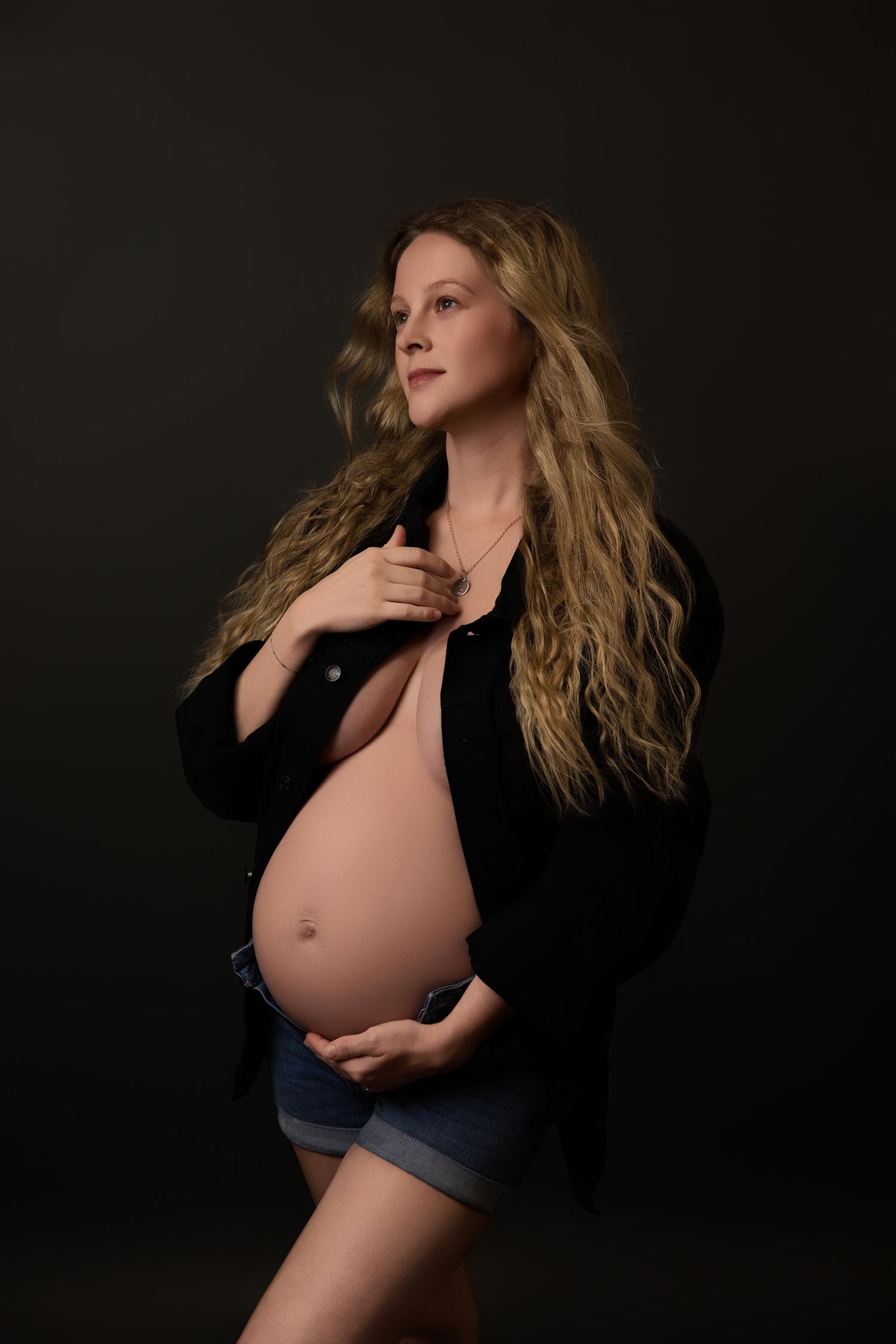 Artistic maternity photography photo with expecting mother in denim shorts and black jacket against dark background.