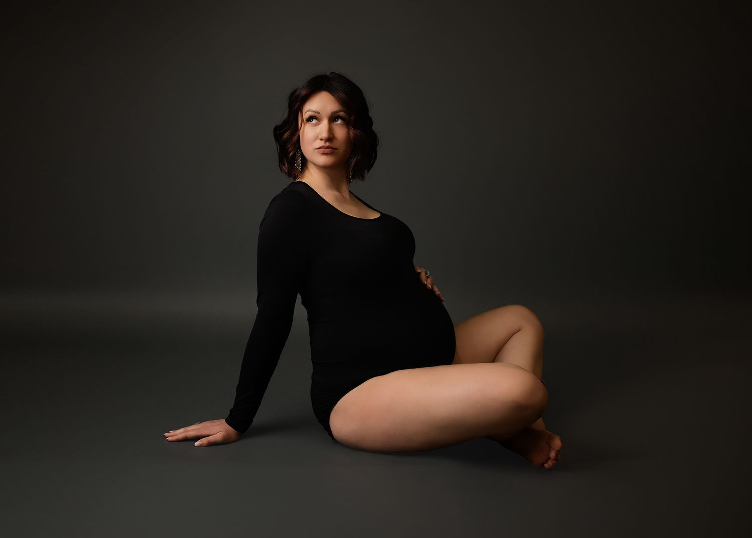 A maternity photographer portrait featuring a figure in a black bodysuit posing elegantly against a dark studio backdrop.