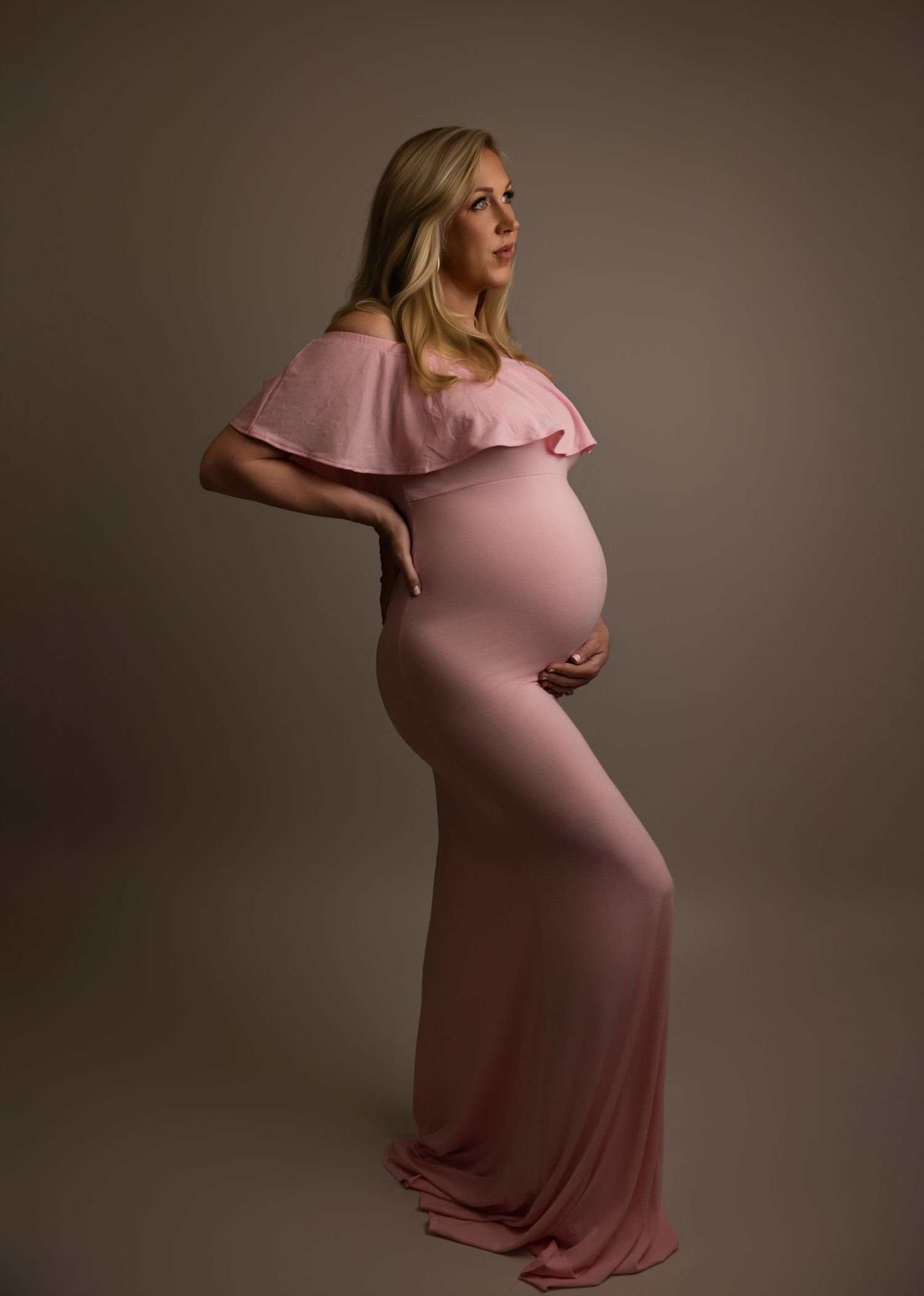 Elegant maternity portrait featuring a flowing pink off-shoulder gown against a neutral brown backdrop.