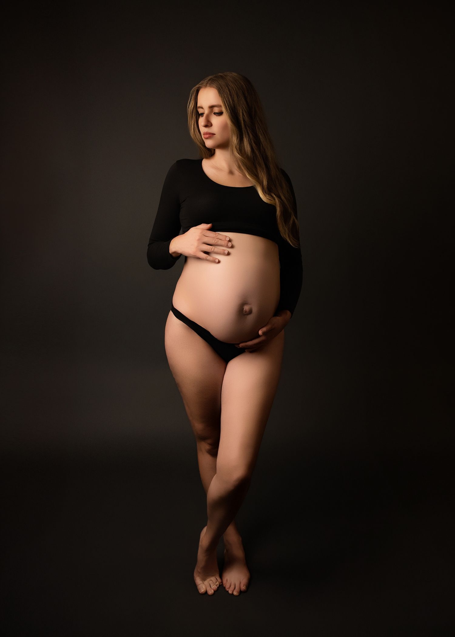 Artistic maternity photographer portrait in dark studio showing pregnant woman in black top and underwear against shadowy background.