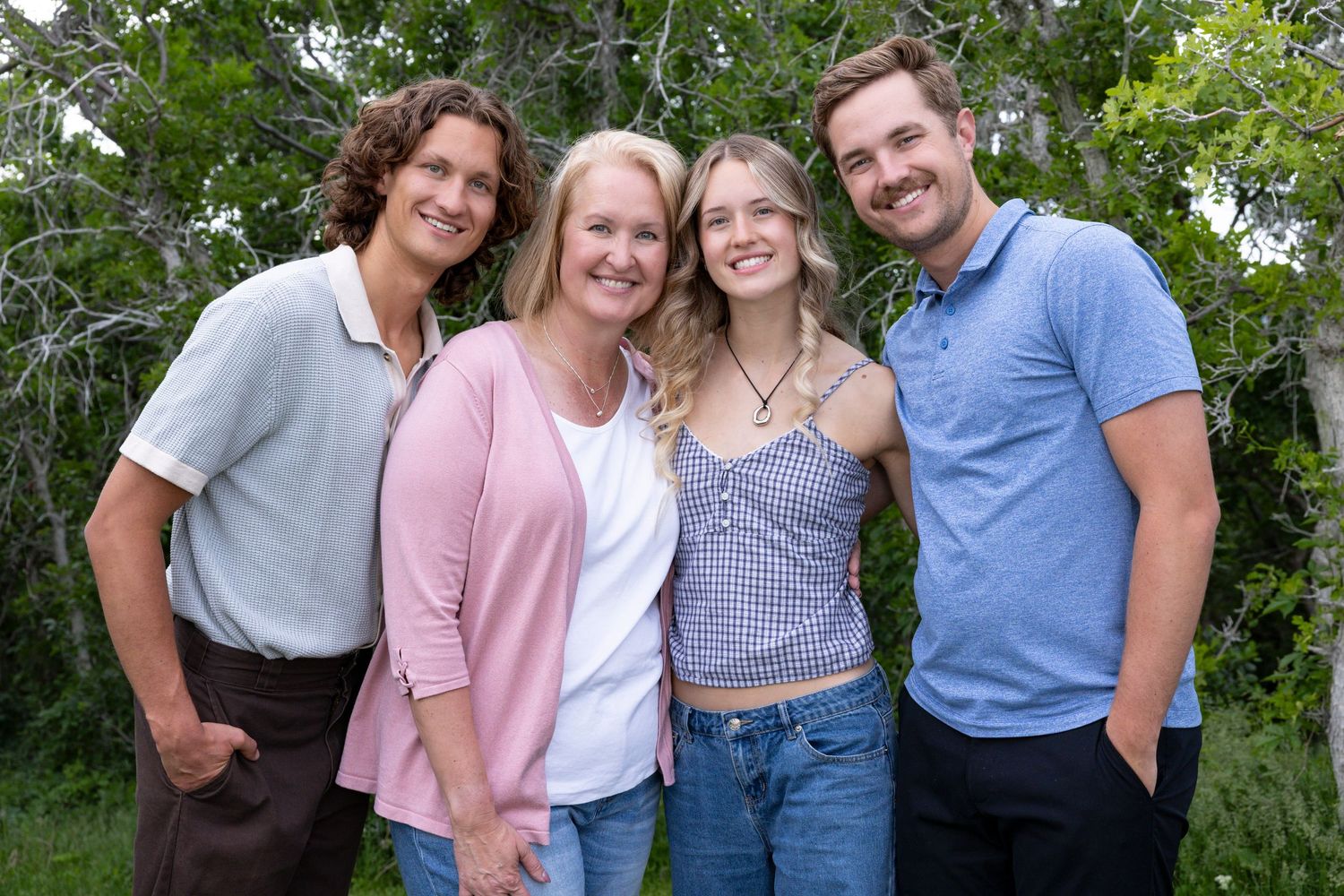 A group of four people standing together outdoors against green foliage wearing casual clothing on a sunny day.