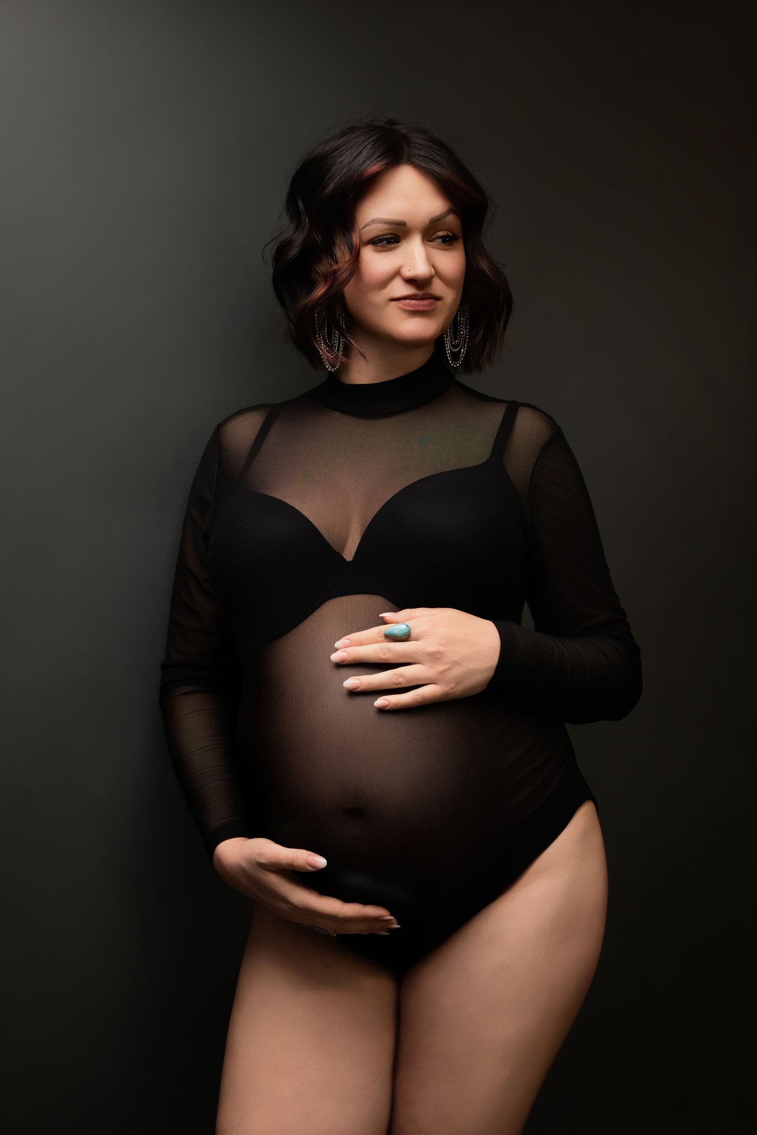 Artistic maternity photographer portrait featuring an expectant mother in a sheer black bodysuit against a dark backdrop.