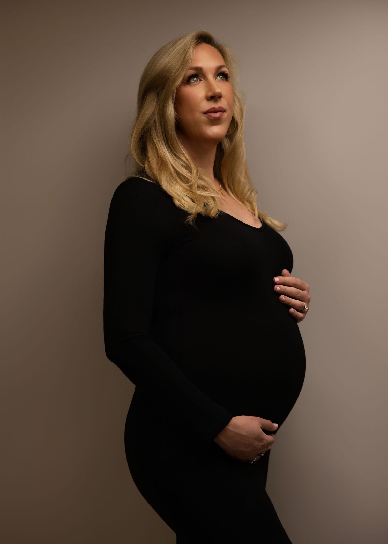 A woman in a black dress poses for a moody maternity photo against a dark brown background.