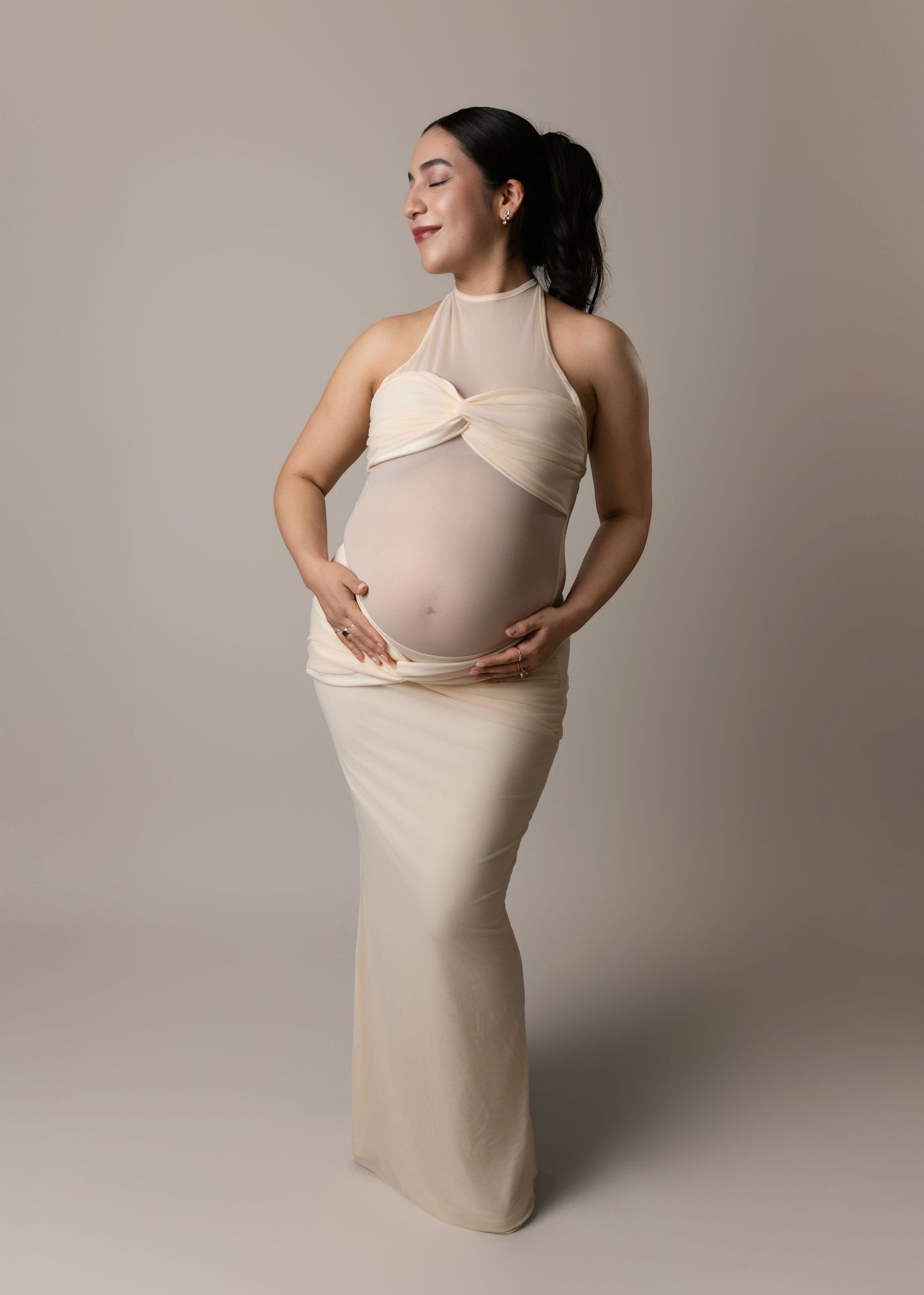 Elegant maternity photo of an expectant mother in a cream-colored halter dress posing against a neutral backdrop.
