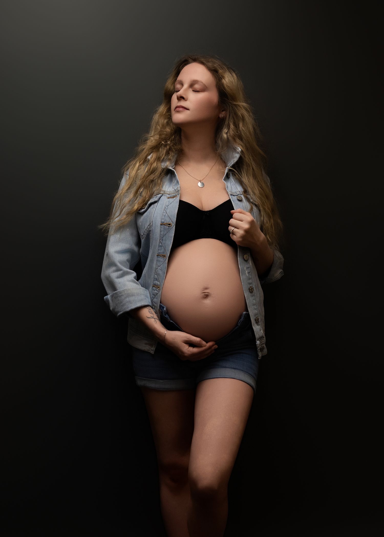 A pregnant woman in denim jacket and shorts poses against a dark background in a moody maternity photographer photo.