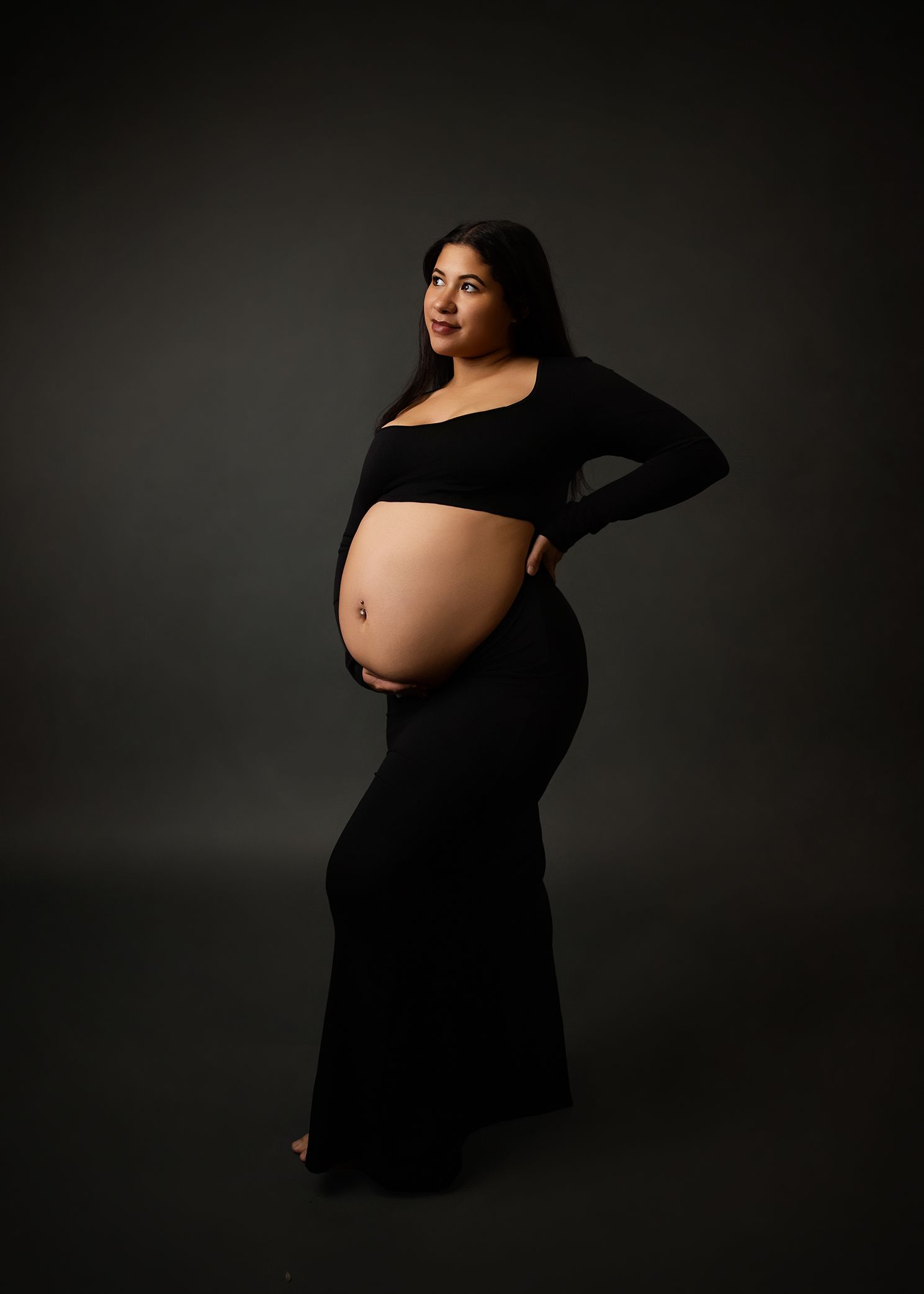 Elegant maternity portrait in black dress against dark background showcasing a pregnant silhouette in dramatic lighting.