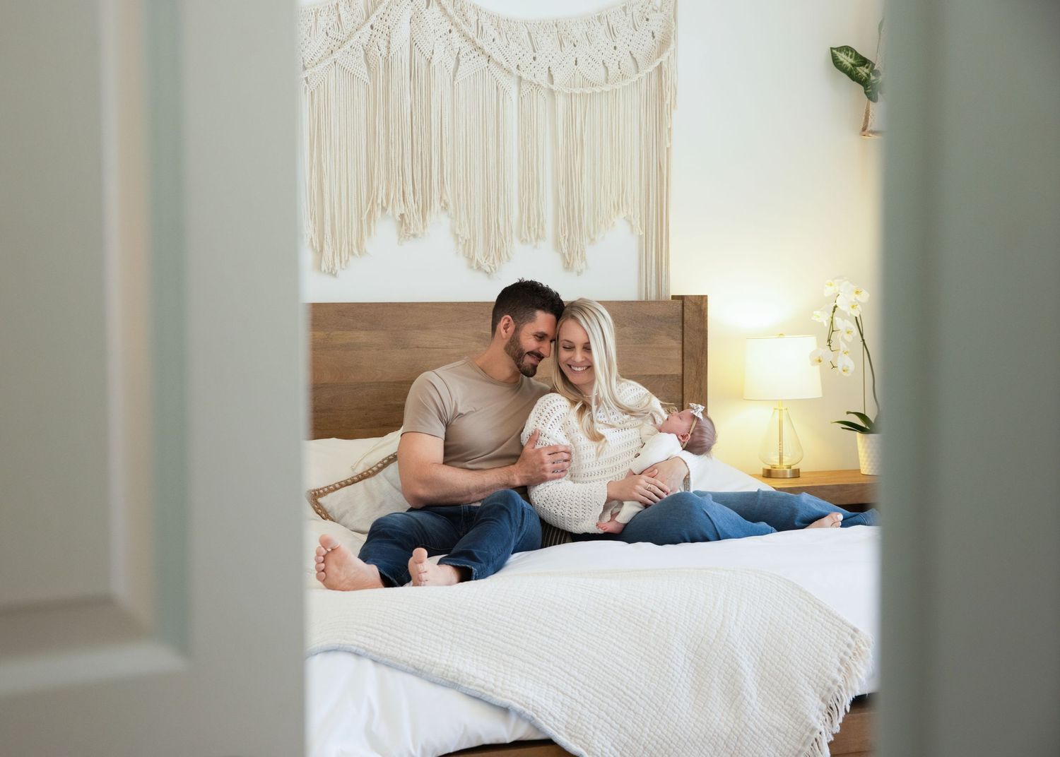 Happy couple sitting together on white bed with macrame wall hanging and lamp in cozy bedroom setting.