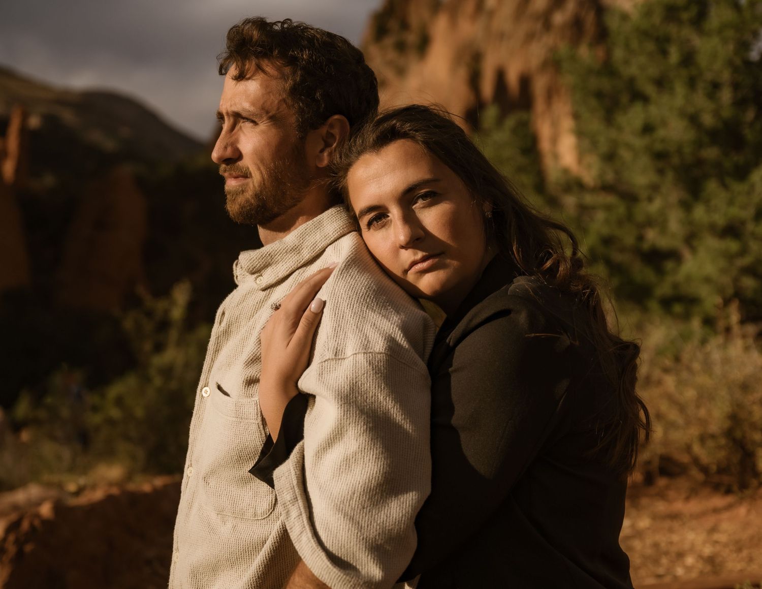 A couple in casual attire poses against a dramatic mountain backdrop at sunset.