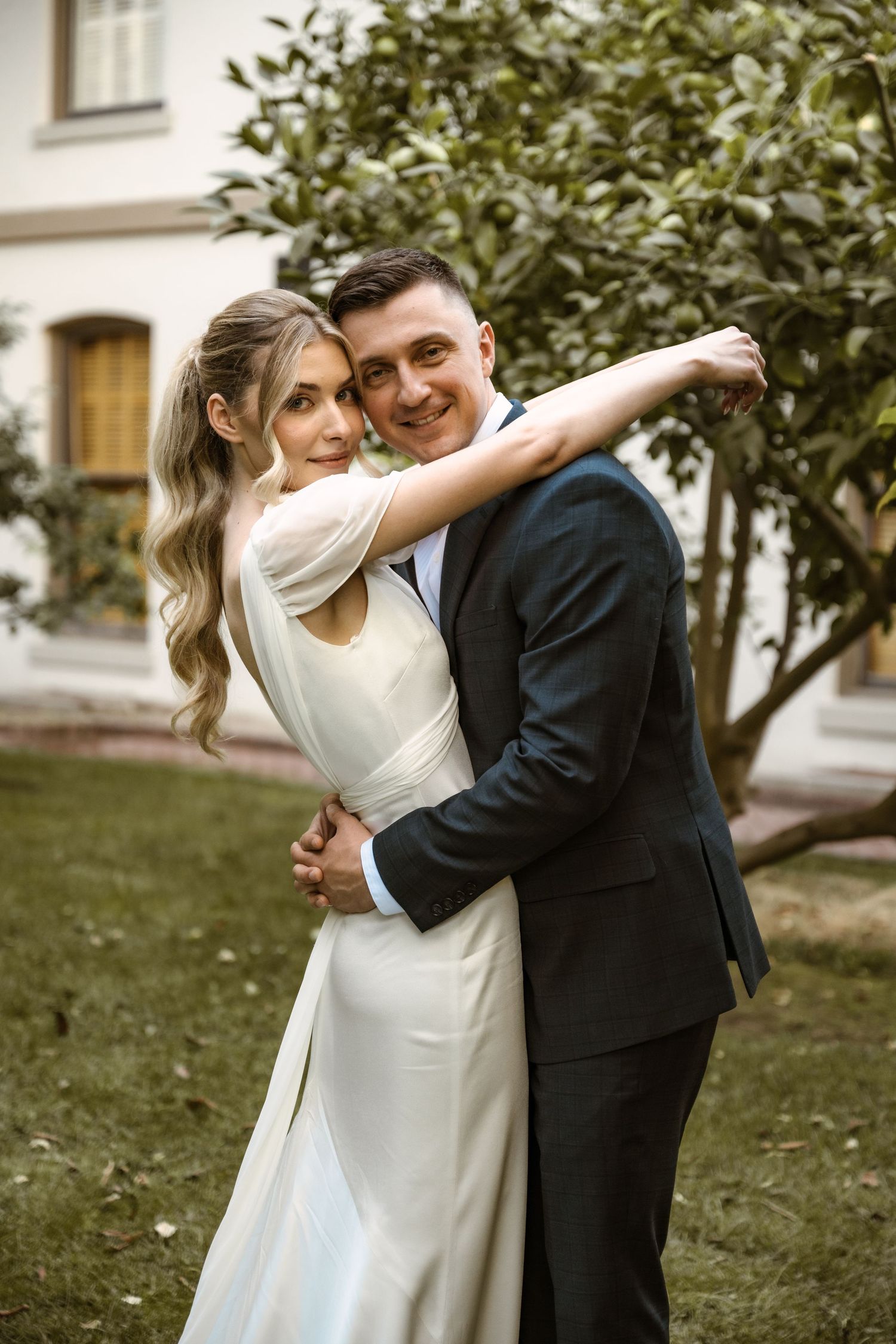 A couple embraces while dancing outdoors near green foliage.