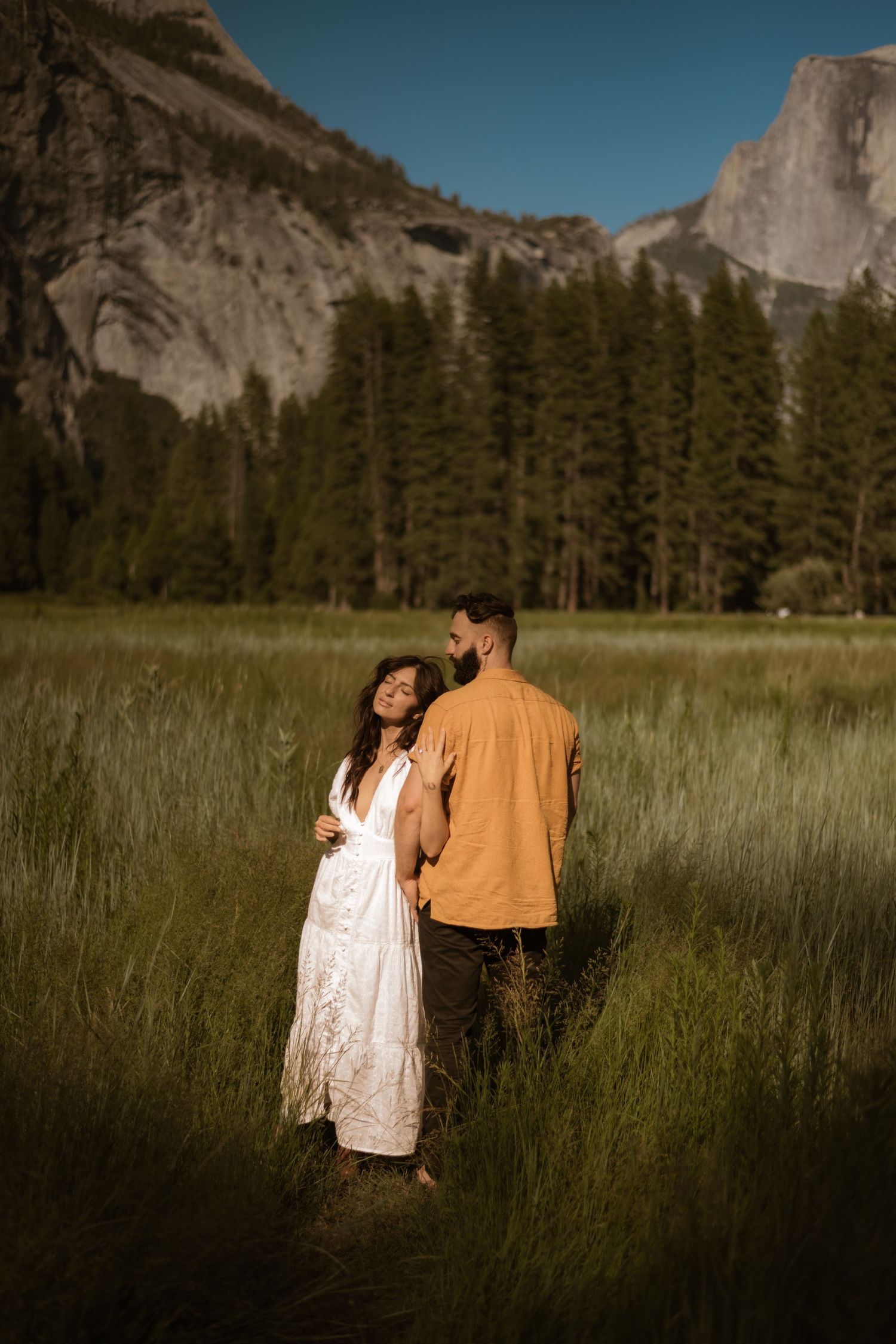 Engagement photos A couple stands in tall grass with Yosemite's Half Dome mountain visible in the background.